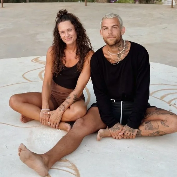 A woman and a man sitting cross-legged on a painted concrete surface outdoors. The woman has long, dark curly hair, and is wearing a black tank top and shorts, with tattoos on her arms. The man has short, gray hair, a beard, and is wearing a black long-sleeve shirt and shorts, also with tattoos on his arms and legs.
