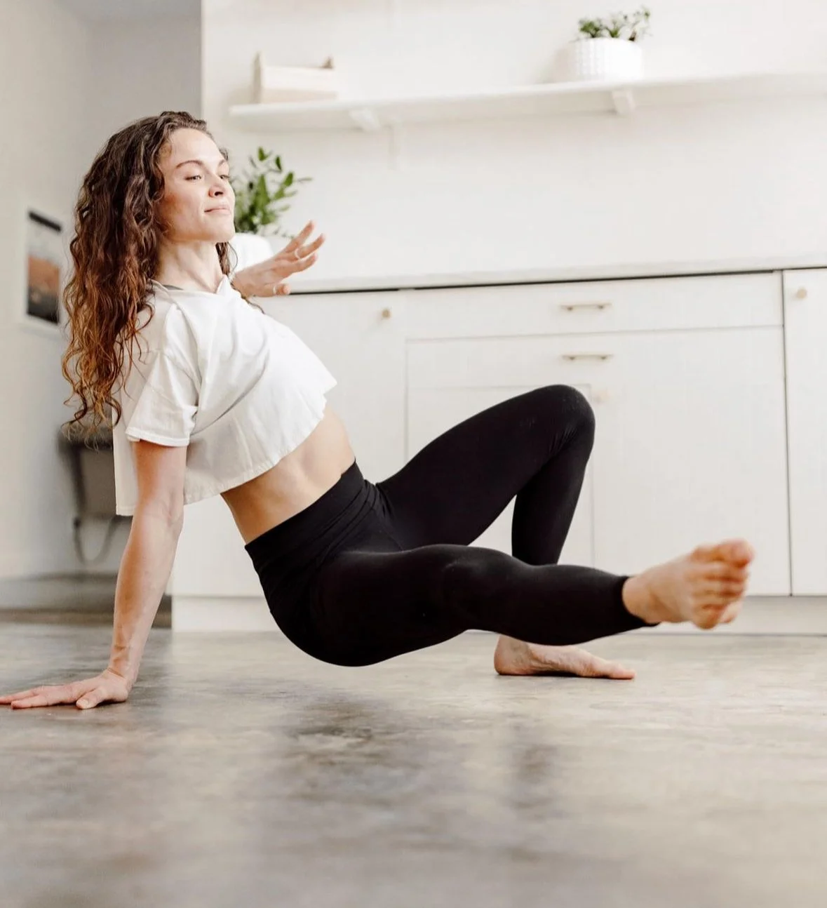 A woman in black leggings and a white crop top practicing yoga or stretching on a wooden floor in a bright room with white cabinets and shelves.