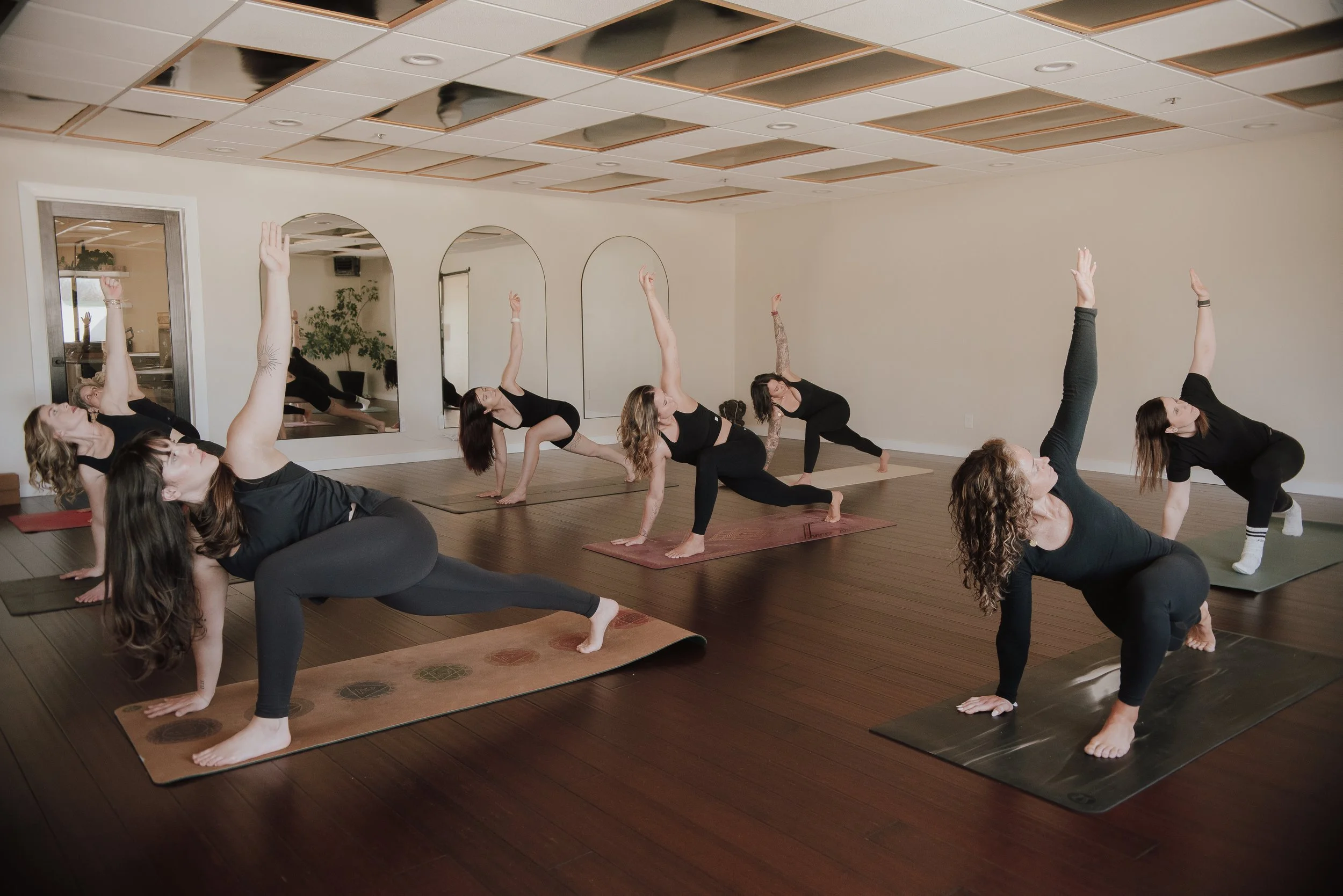 Group of women participating in a yoga class in a studio, practicing yoga poses on mats with mirrors on the wall.