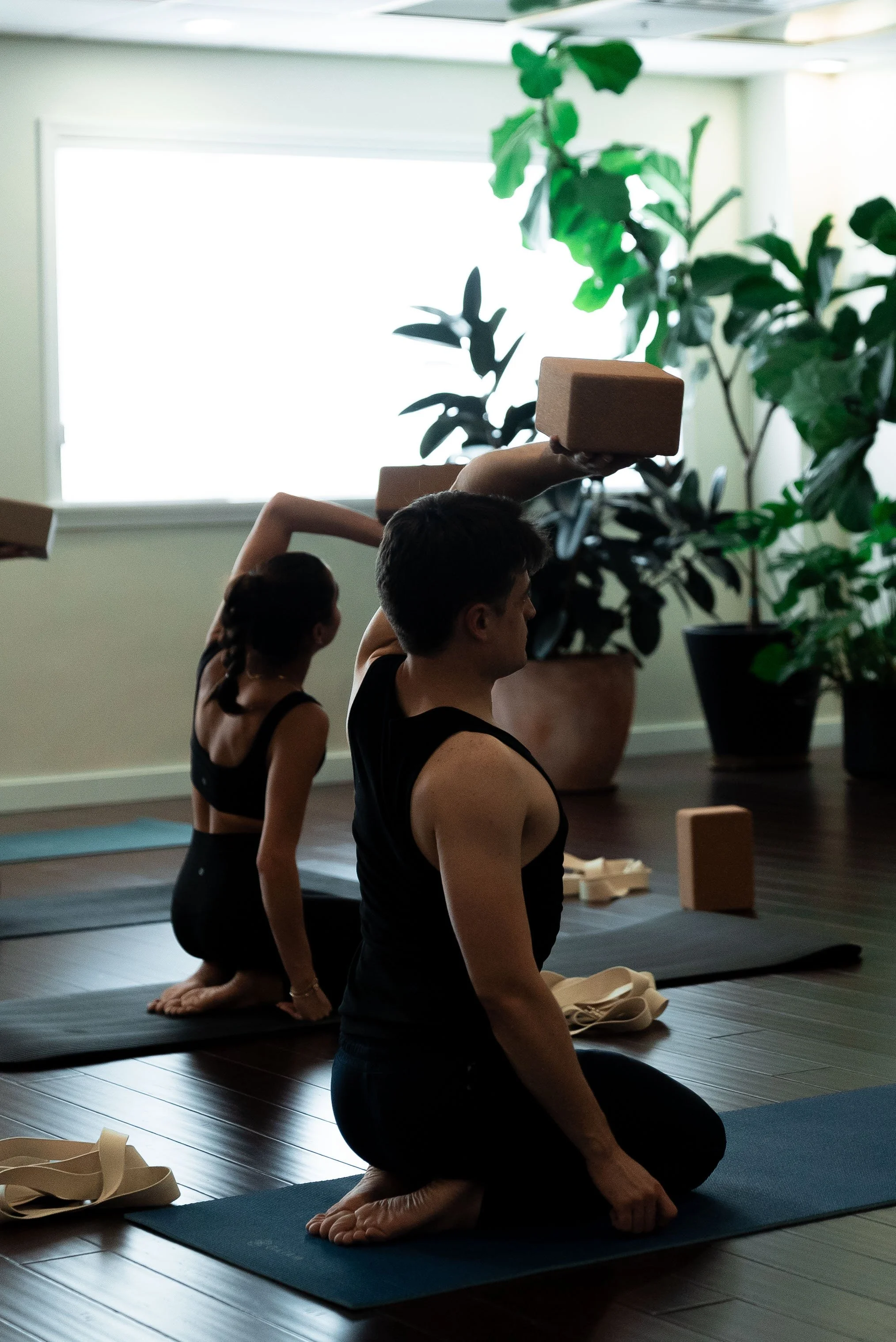 Three people participating in a yoga or meditation class indoors on yoga mats, with large potted plants nearby and natural light from a window.
