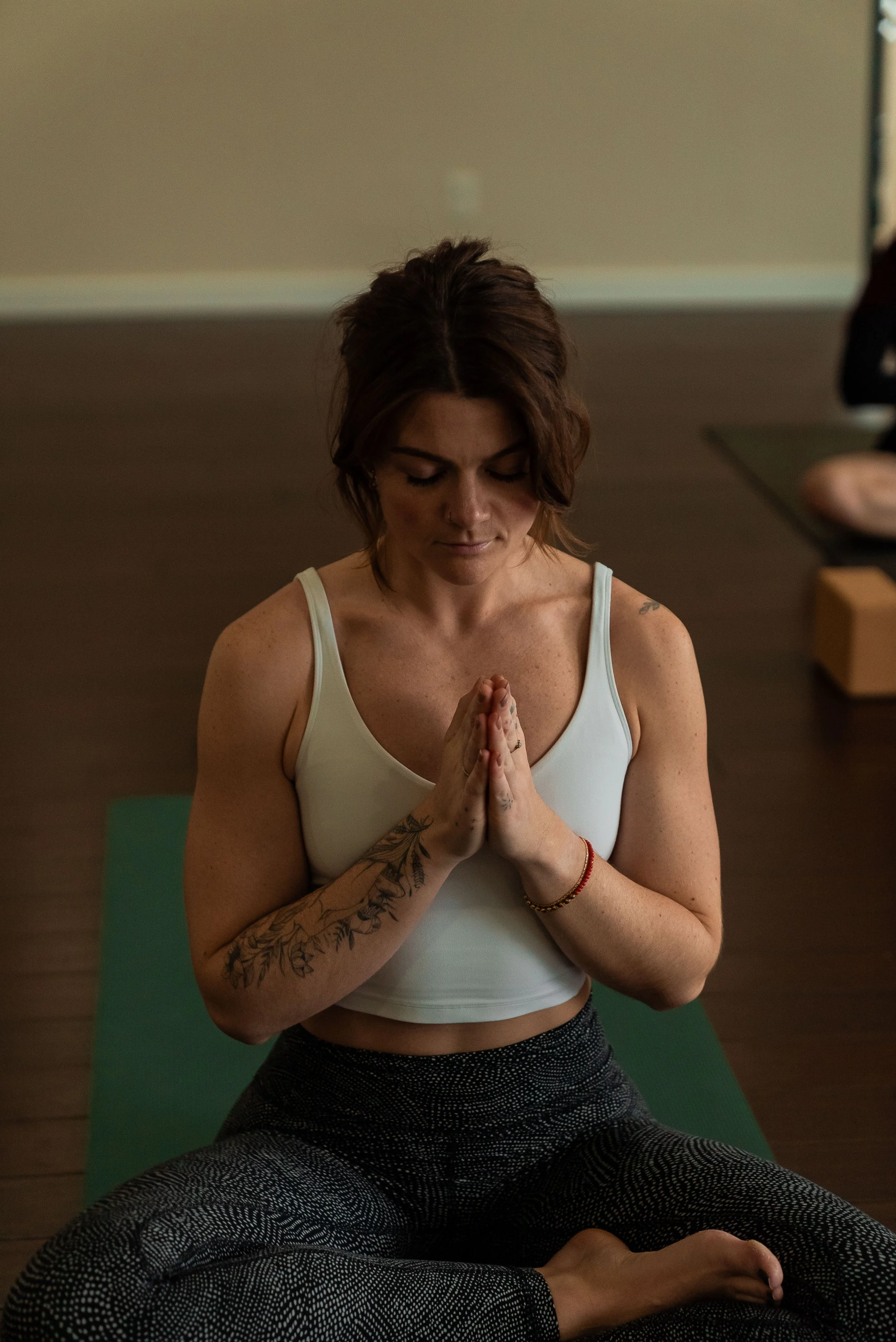 A woman practicing yoga indoors, seated cross-legged with hands pressed together in a prayer position, eyes closed in meditation.