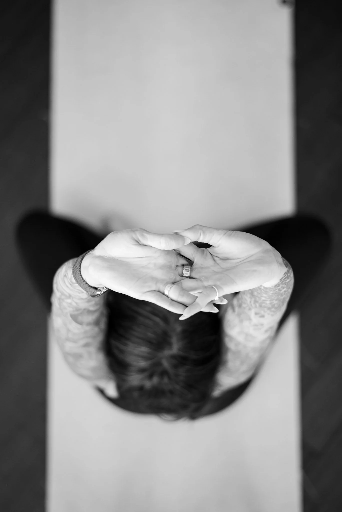 Black and white aerial view of a woman with dark hair and tattoos, practicing meditation or yoga with hands in a prayer position on a mat.