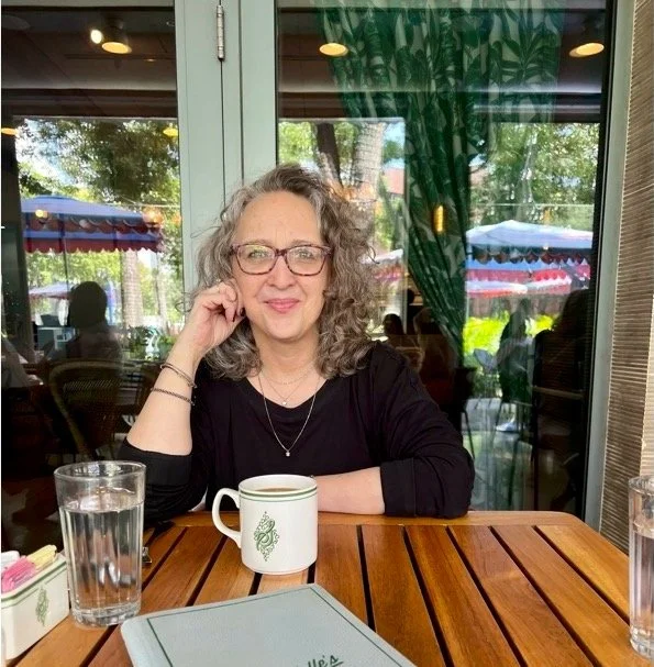 A woman with curly gray hair and glasses sitting at a wooden table in a restaurant or cafe, smiling with her head resting on her hand, with a white coffee mug, a glass of water, and a menu in front of her. The background shows large windows with green curtains and outdoor patio umbrellas.