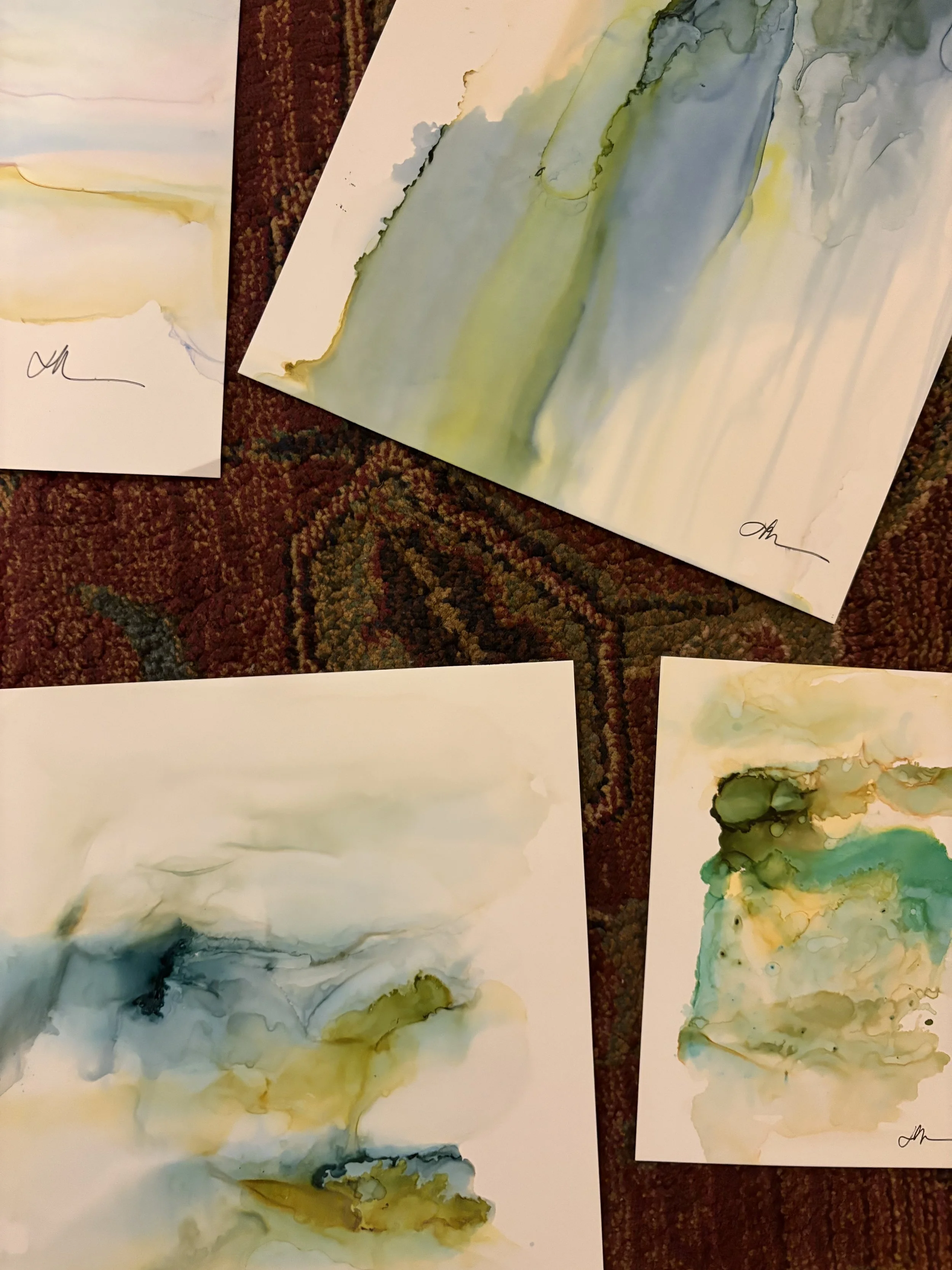 Four sheets of abstract watercolor paintings in shades of green, blue, yellow, and beige lying on a red patterned carpet.