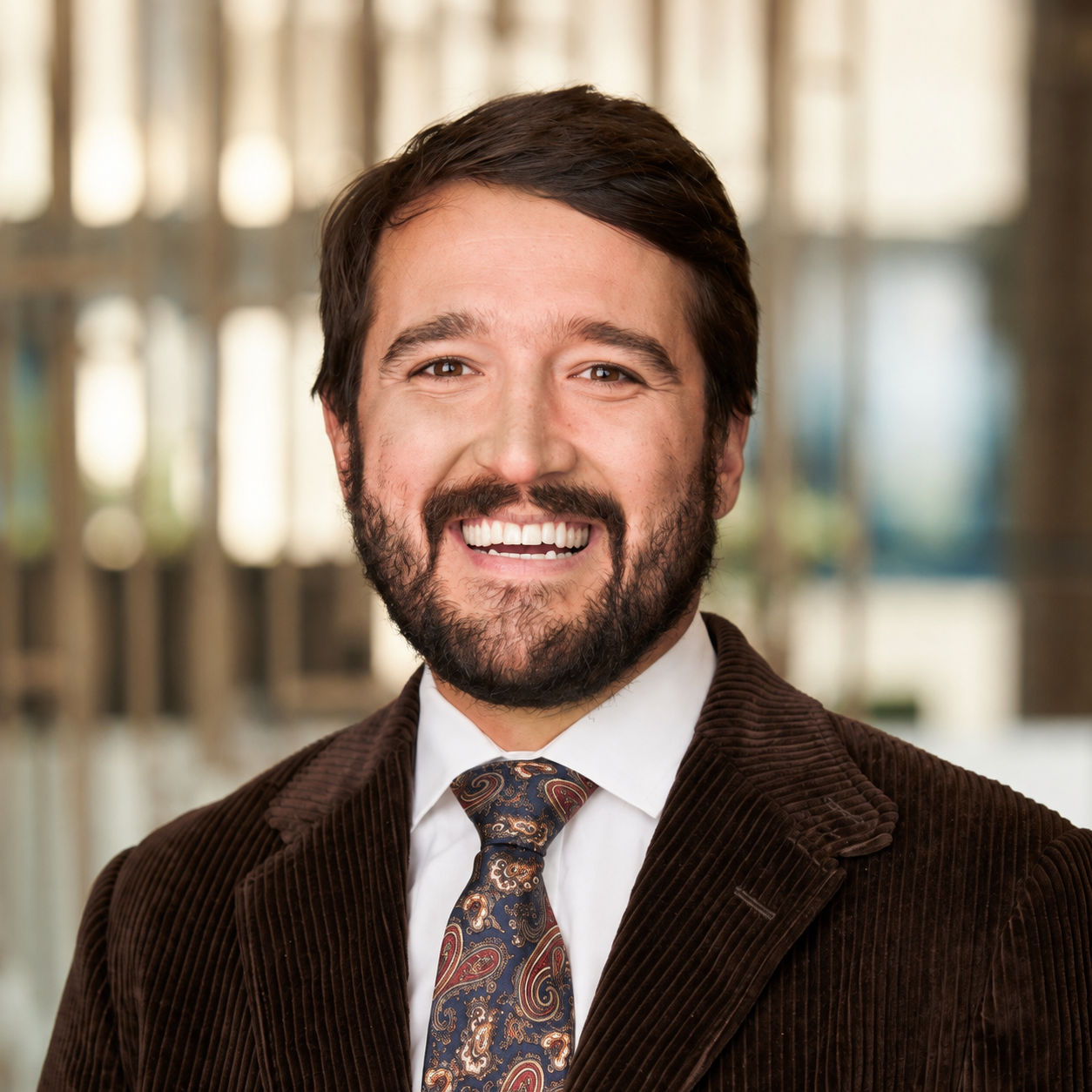 A smiling man with dark hair, a beard, wearing a brown blazer, white shirt, and a patterned tie, standing outdoors with blurred background.