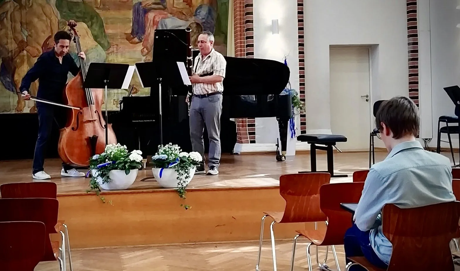 Zwei Musiker auf einer Bühne während einer Probe; einer spielt Kontrabass, der andere spielt Fagott; im Hintergrund ein Klavier und eine Wand mit Wandbild; im Vordergrund sitzt der Komponist und liest die Partitur.