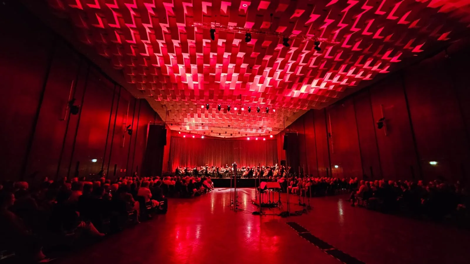 Ein dunkler Konzertsaal mit roter Beleuchtung, auf der Bühne spielen Orchestermitglieder, das Publikum sitzt im Saal.