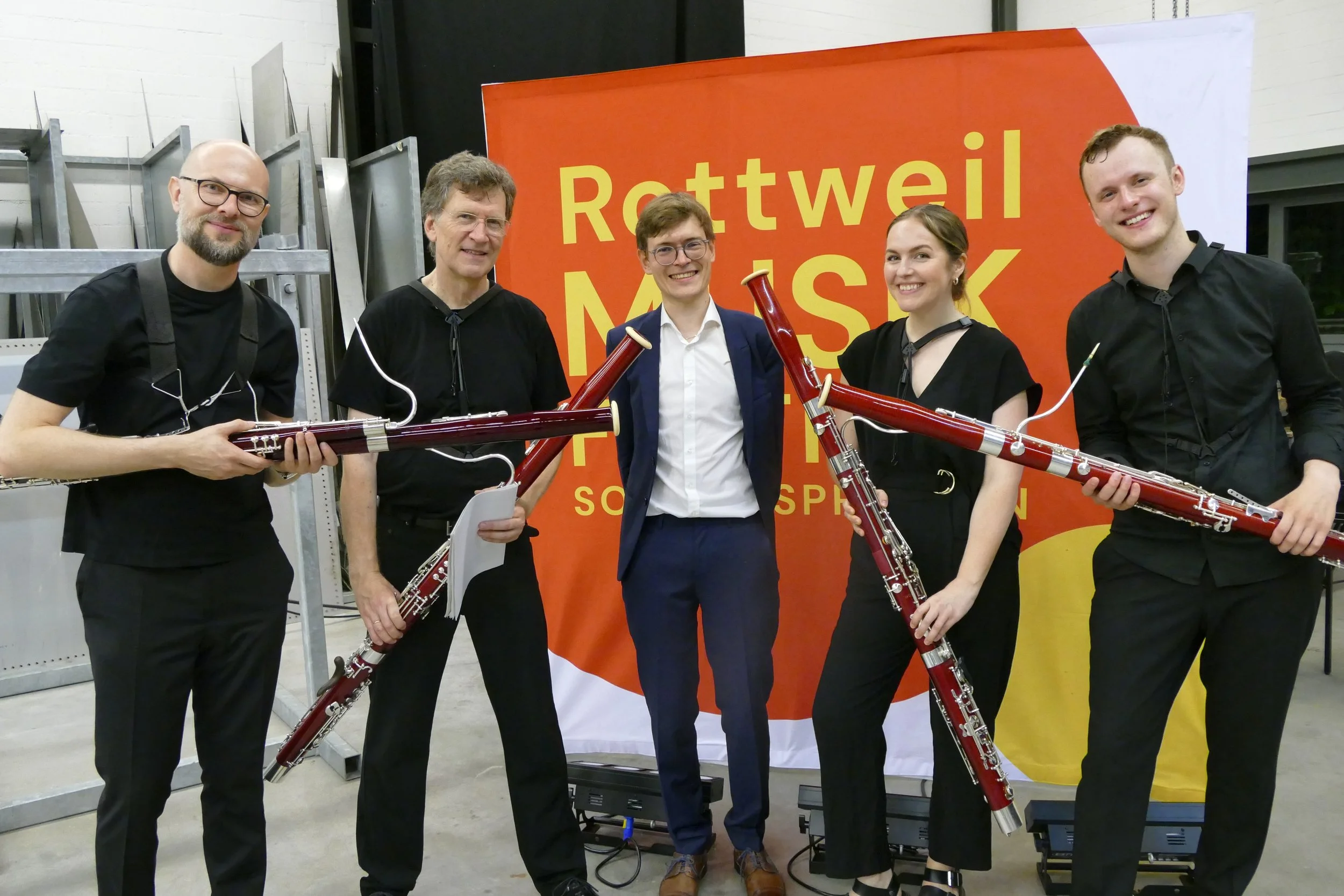 Fünf Menschen stehen nebeneinander vor einem orangefarbenen Hintergrund mit dem Schriftzug "Rottweil Musik". Sie halten Fagotte und lächeln in die Kamera. Die Gruppe ist beim Musikfestival Sommersprossen in Rottweil.