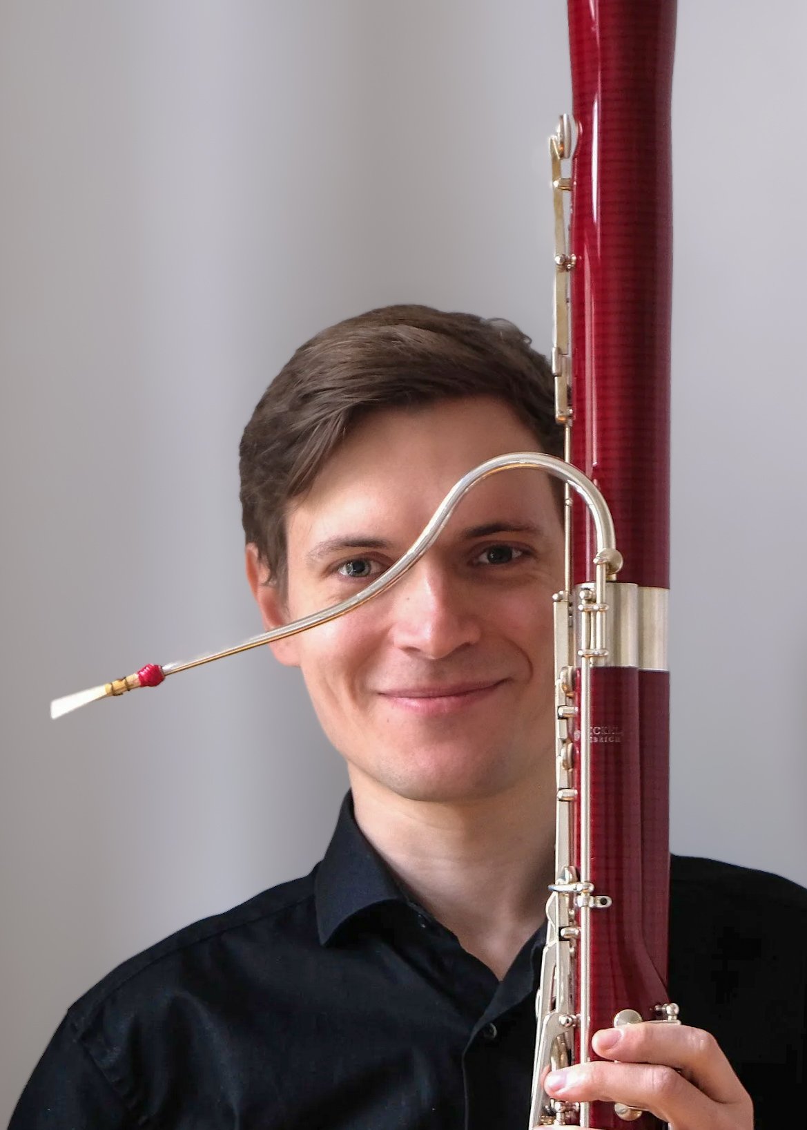 Ein Mann hält ein Fagott vor seinem Gesicht, die ihn teilweise verdeckt. Er trägt ein schwarzes Hemd und hat braune, kurze Haare. Die Hintergrund ist grau.