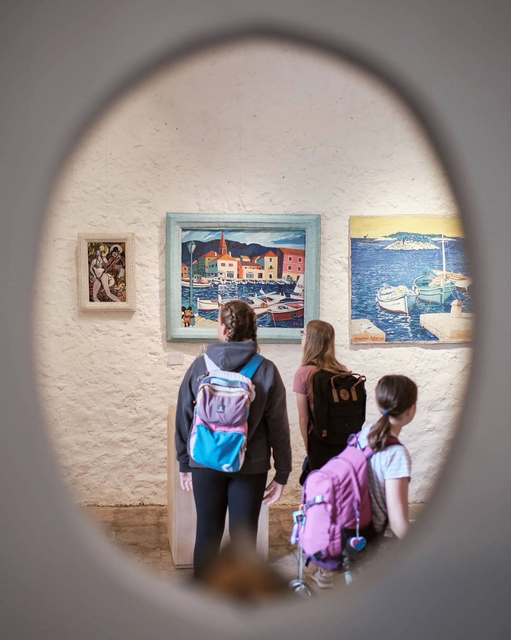 Three children view paintings of a harbor scene in an art gallery, framed by an oval-shaped opening in the wall.