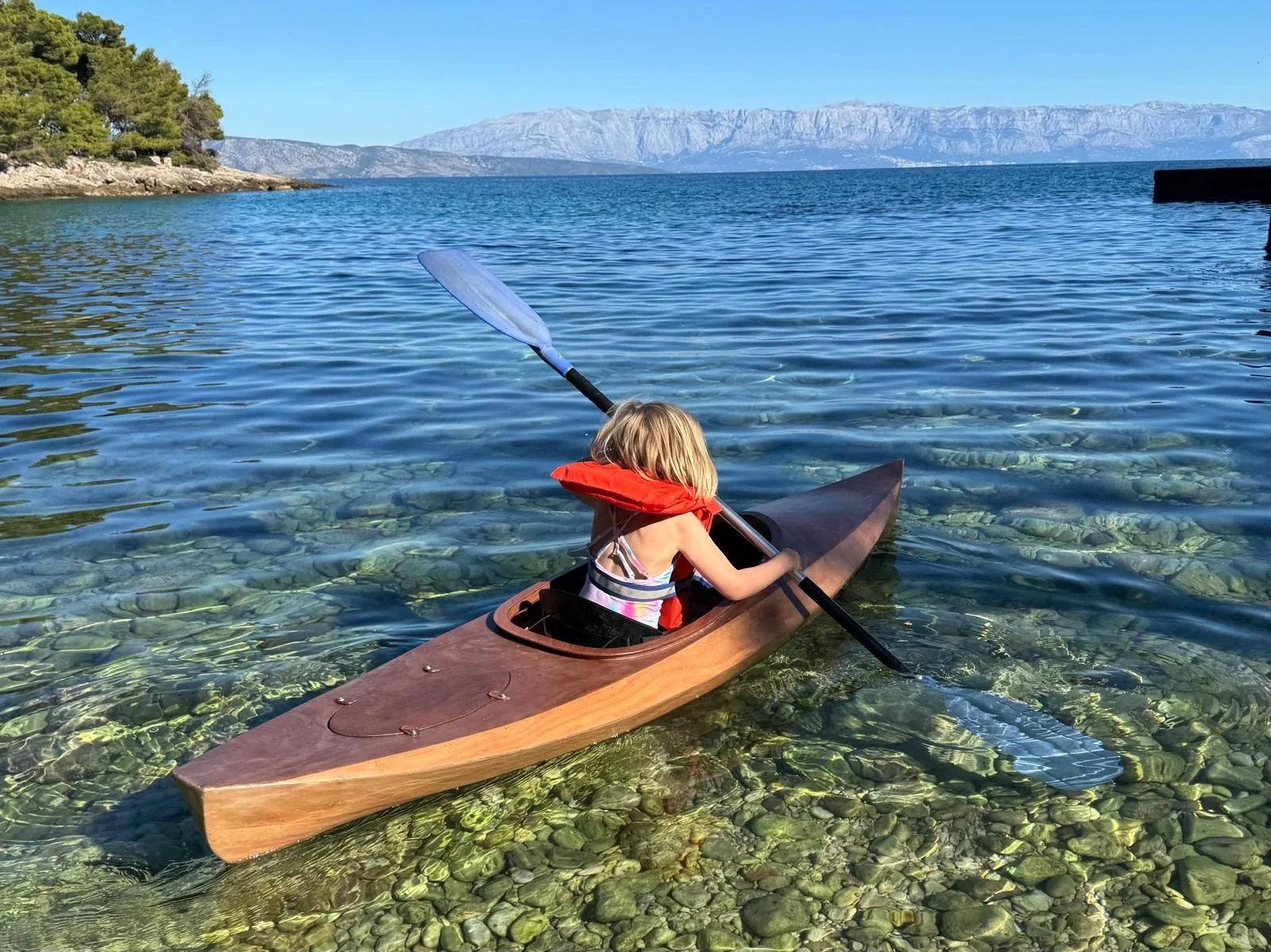 A young girl with blonde hair in a kayak on a clear body of water, with mountains in the background and trees on the shore.