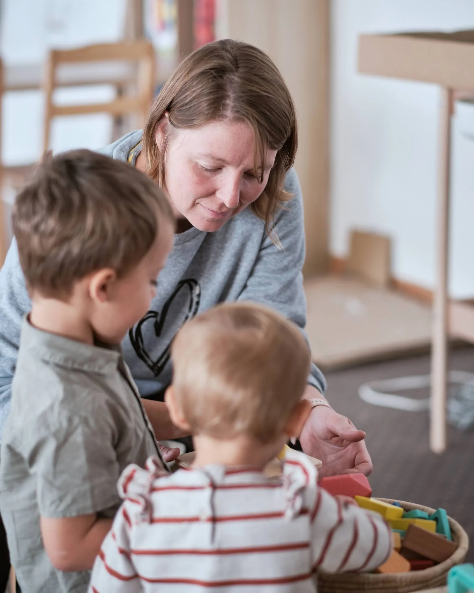 A woman helps two young boys with a toy activity in an indoor setting, likely a classroom or play area.