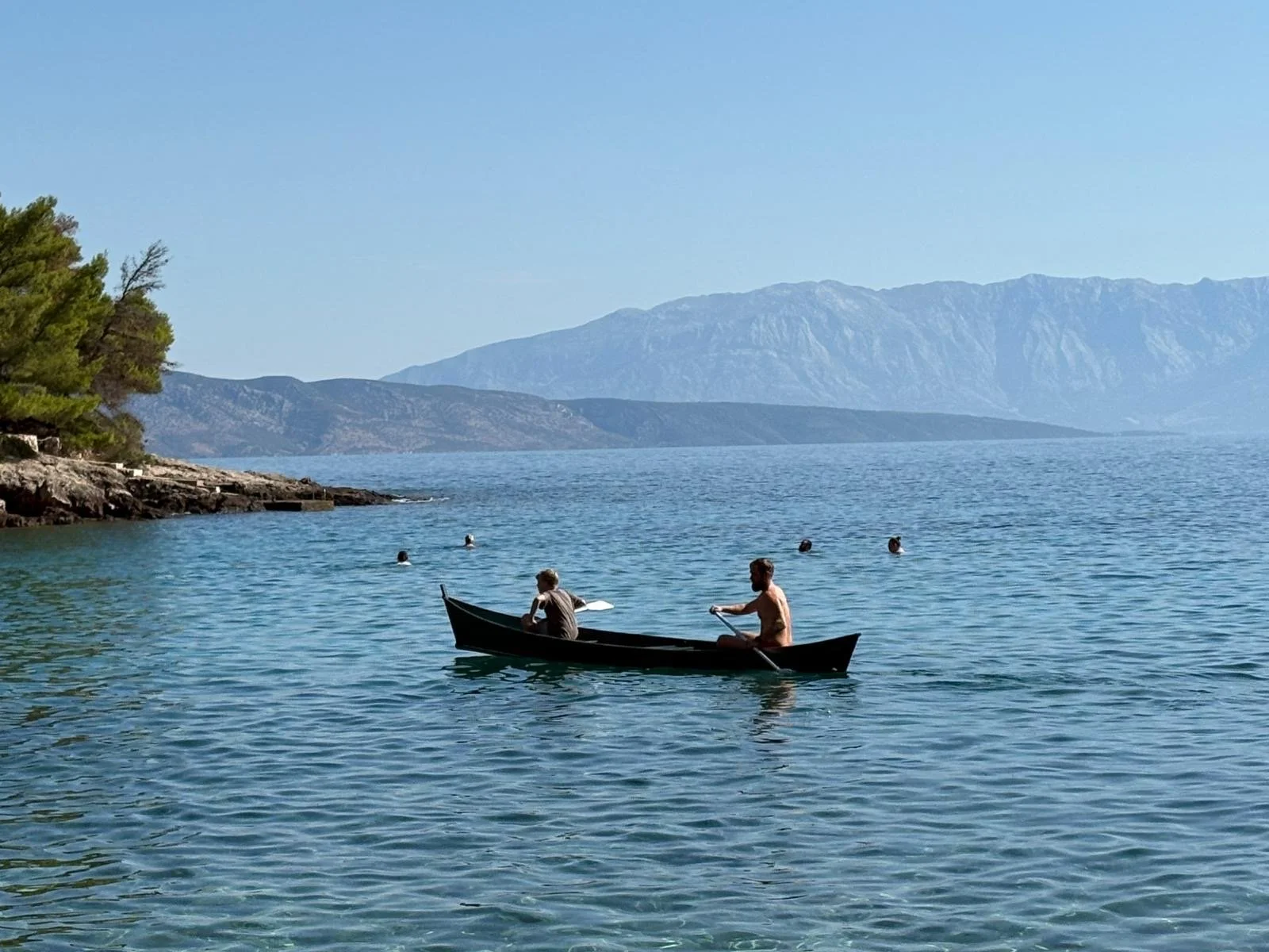 Two men in a black boat on a calm blue lake, with three floating swimmers nearby, surrounded by green trees on the left and mountain ranges in the background under a clear blue sky.