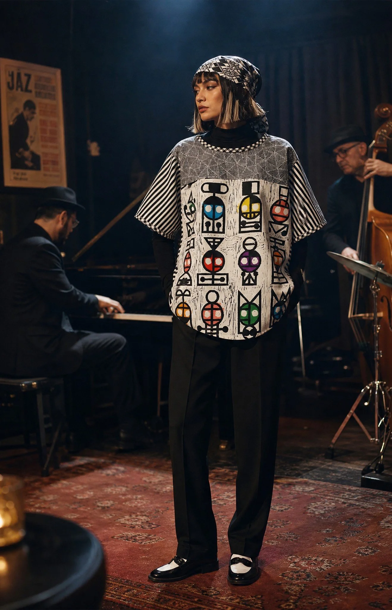 A woman standing on a patterned rug in front of jazz musicians playing piano and upright bass, wearing a graphic top with abstract designs, black pants, and two-tone shoes, with a jazz poster visible in the background.