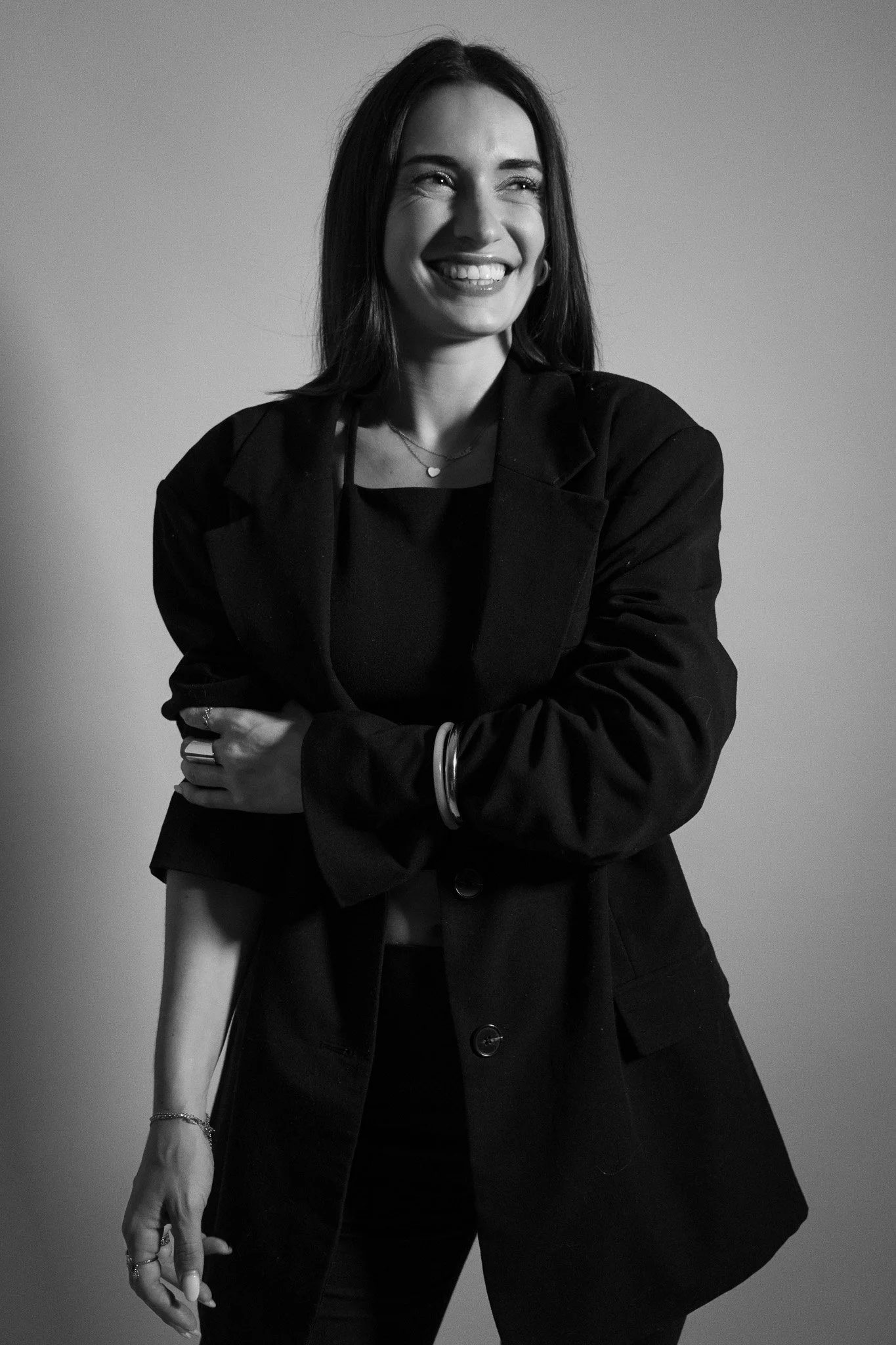 Une femme souriante portant une veste noire et des accessoires, debout contre un fond gris.