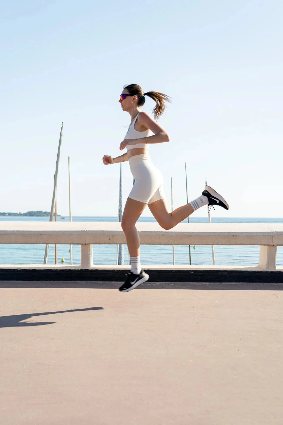 Une femme en tenue de sport court en courant le long d'une promenade en bord de mer, portant des lunettes de soleil, une veste crop top blanche, un short blanc, des baskets noires, et des chaussettes rayées.