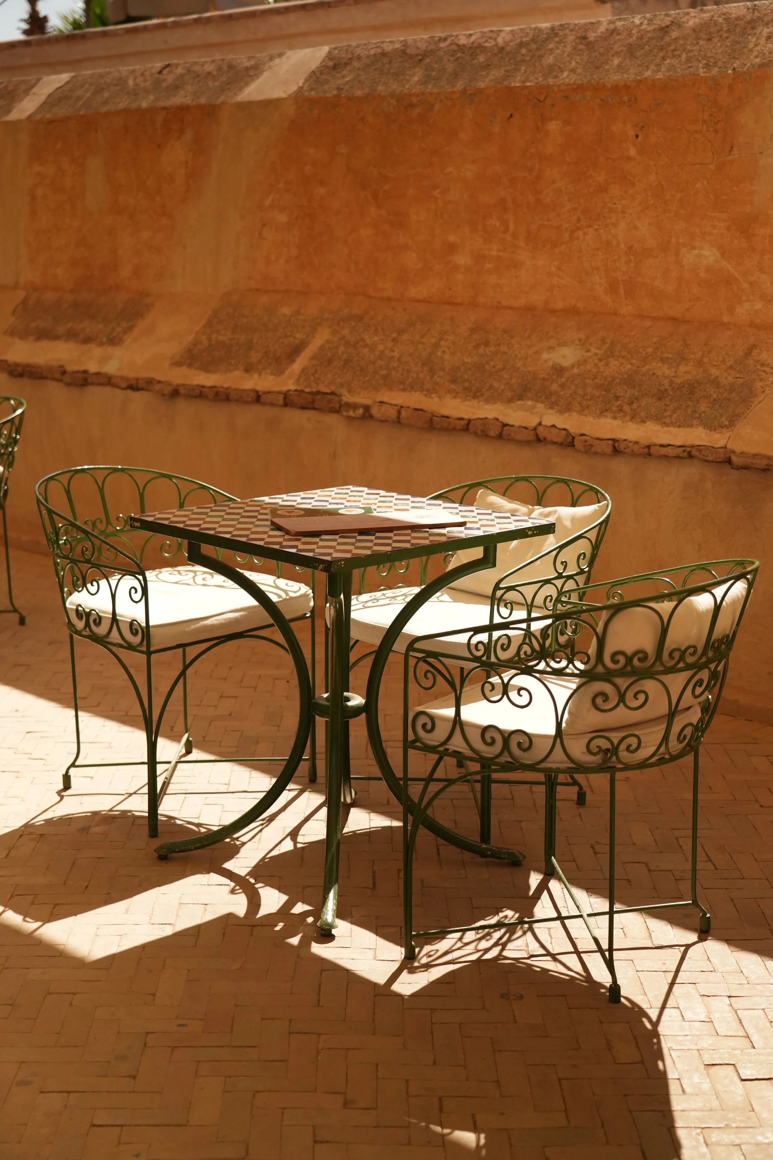 Une table en métal avec un plateau en mosaïque, entourée de quatre chaises en métal à dossiers en fer forgé, sur une terrasse en brique, avec un mur en terre cuite en arrière-plan.