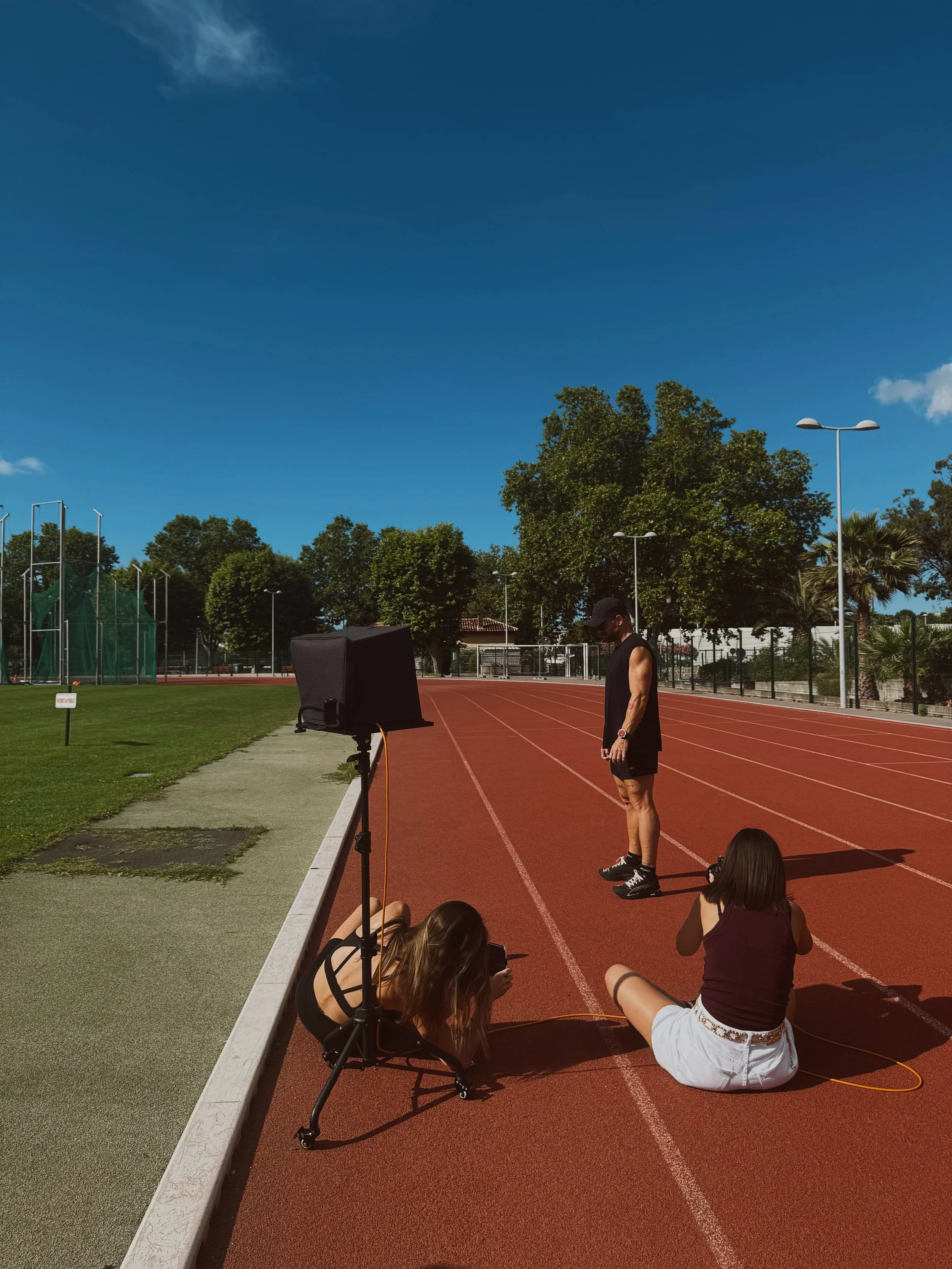 Une séance de prise de vue photographique sur une piste d'athlétisme en plein air avec plusieurs personnes, y compris une femme pointant vers un écran de présentation, et une équipe technique utilisant des équipements photographiques, sous un ciel bleu avec quelques nuages.