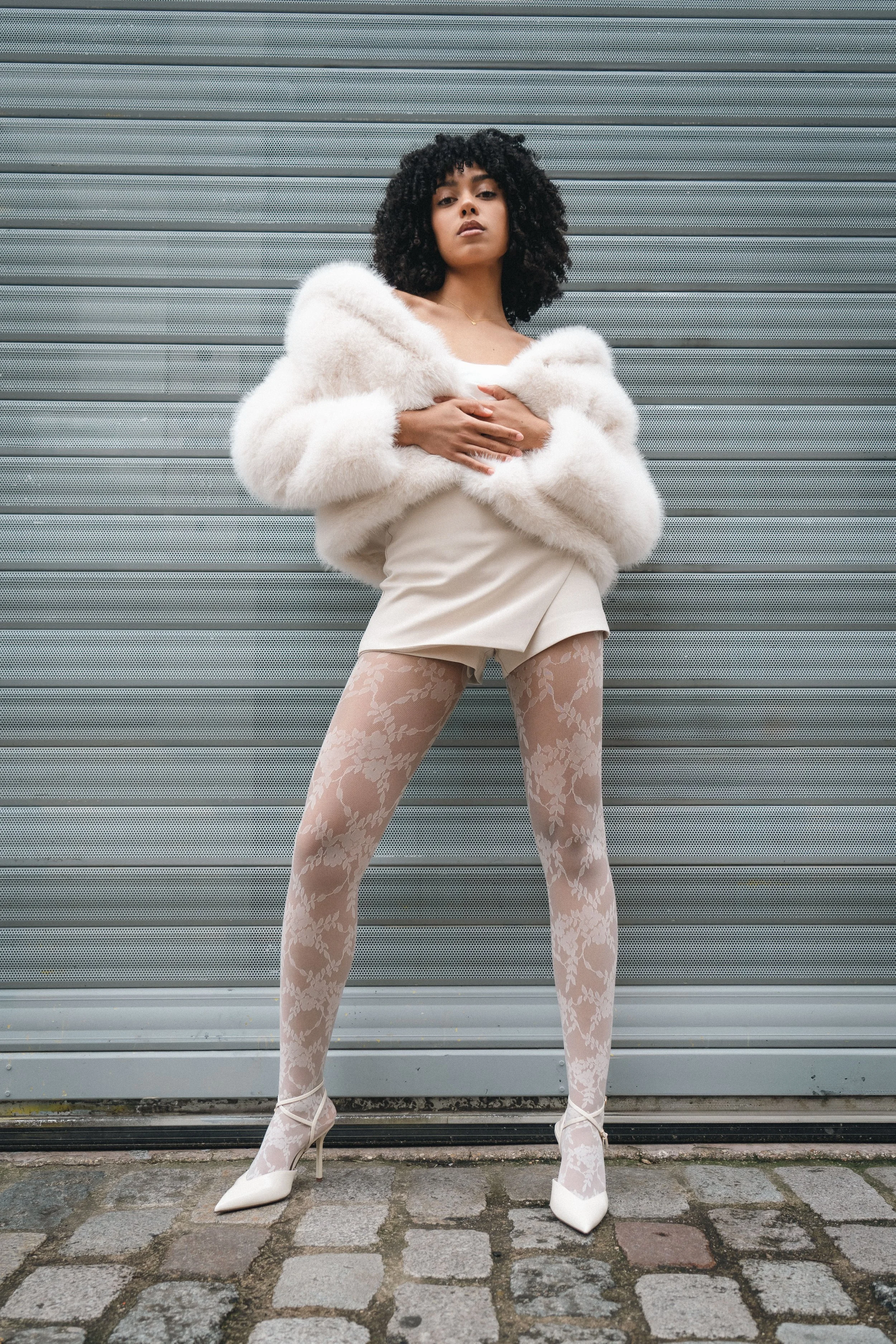Une femme pose devant un mur en métal, portant une robe courte, des collants et des chaussures à talons blancs, avec une manteau en fourrure blanc.