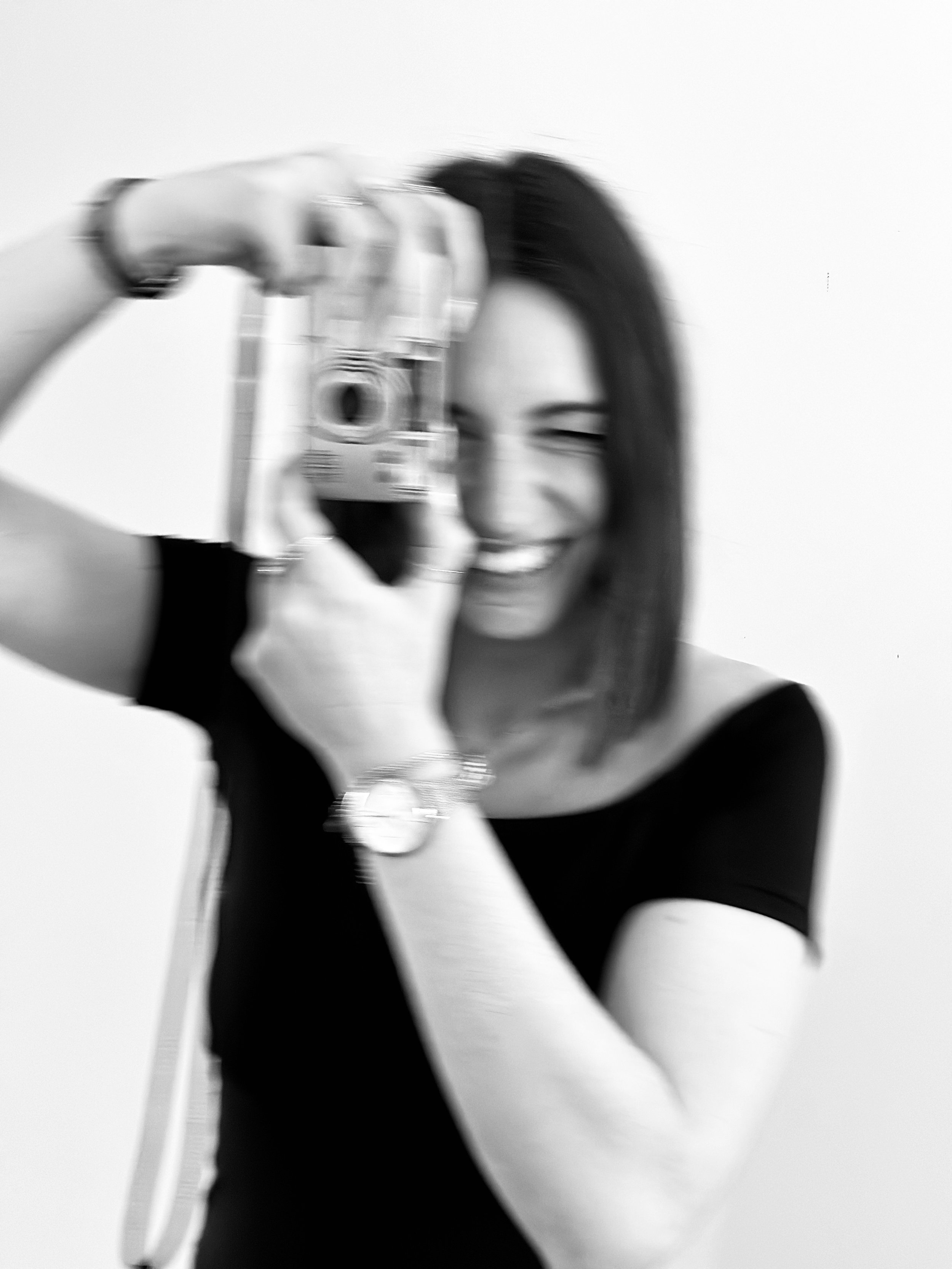 Une femme souriante en noir et blanc, tenant un appareil photo prêt à prendre une photo.