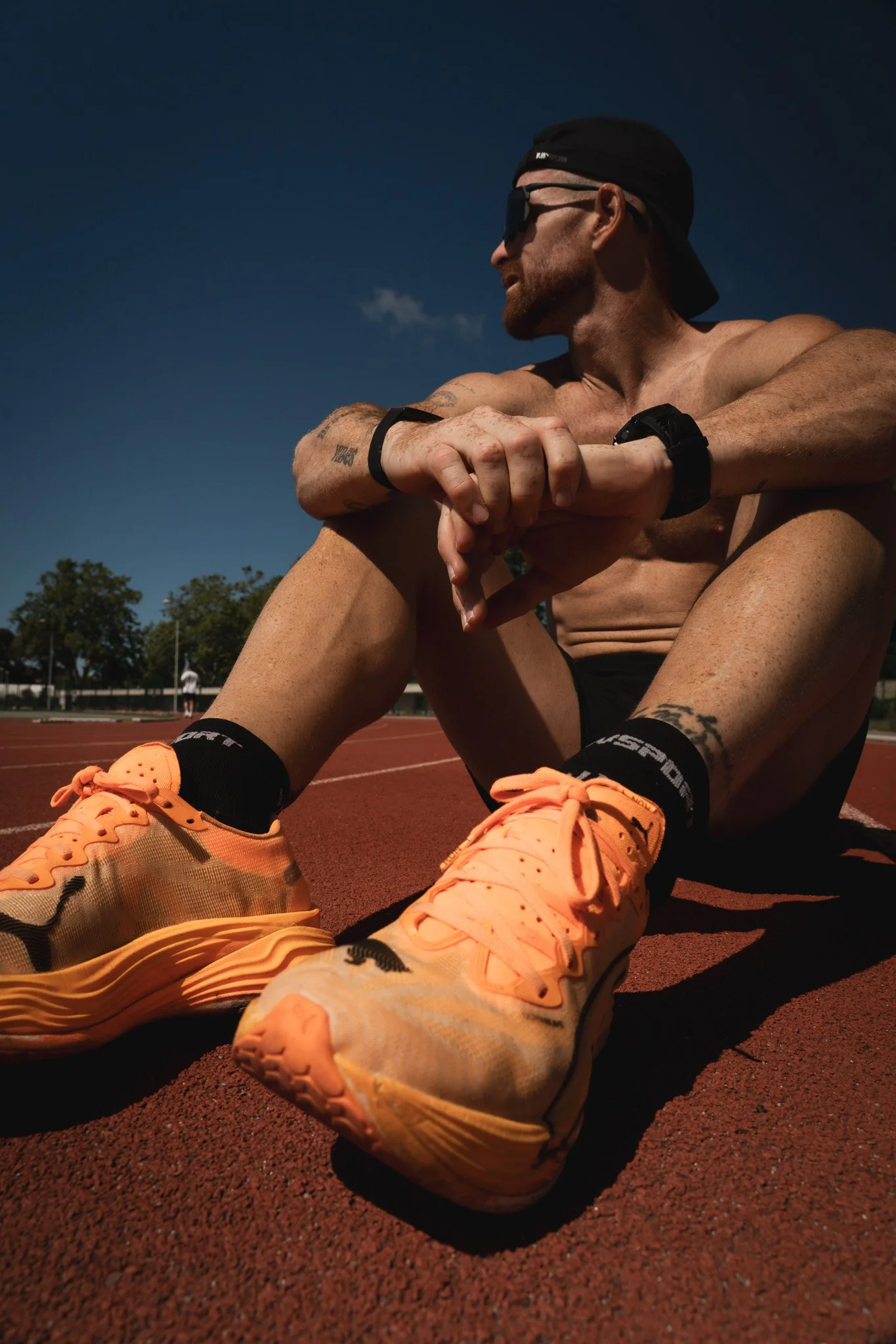 Un homme torse nu, portant une capuche noire, des lunettes de soleil et un bracelet noir, assis sur une piste d'athlétisme, portant des chaussures de course orange, regardant vers la droite, avec un ciel clair en arrière-plan.