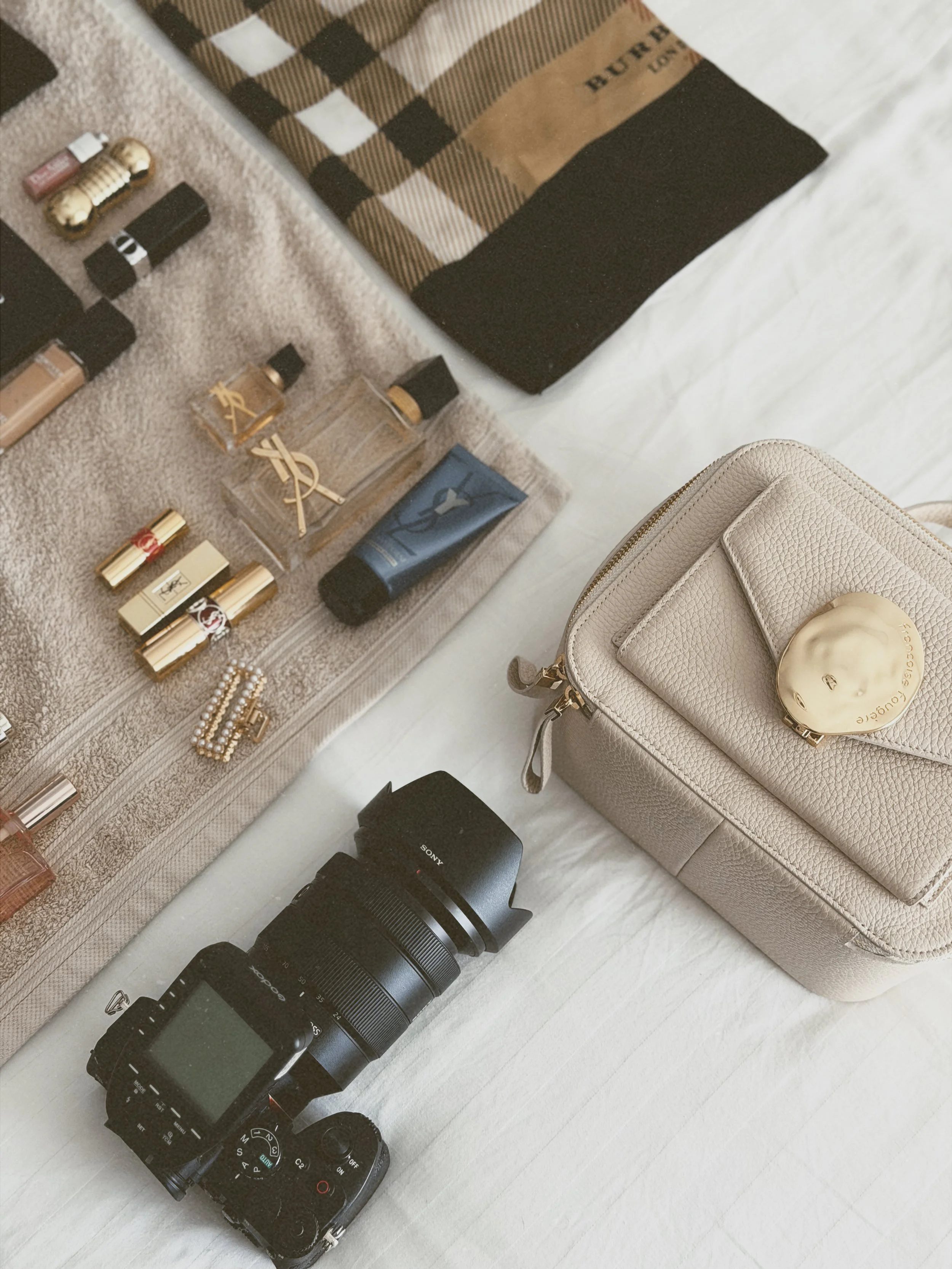 Collection de produits de beauté et accessoires, y compris des tubes de maquillage, un parfum YSL, des bijoux, un appareil photo Sony, un objectif, et un sac beige avec un miroir rond.