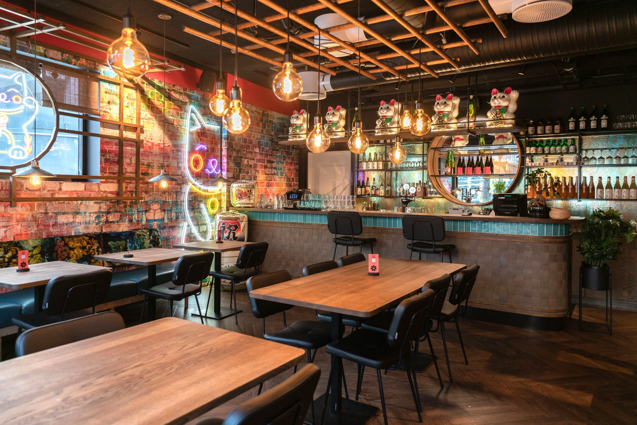 Interiør av en bar eller restaurant med trebord, svarte stoler, en bar med flasker og glass, dekorative figurer av heldigste kattefigurer, neonbord og elegante lyspærer.