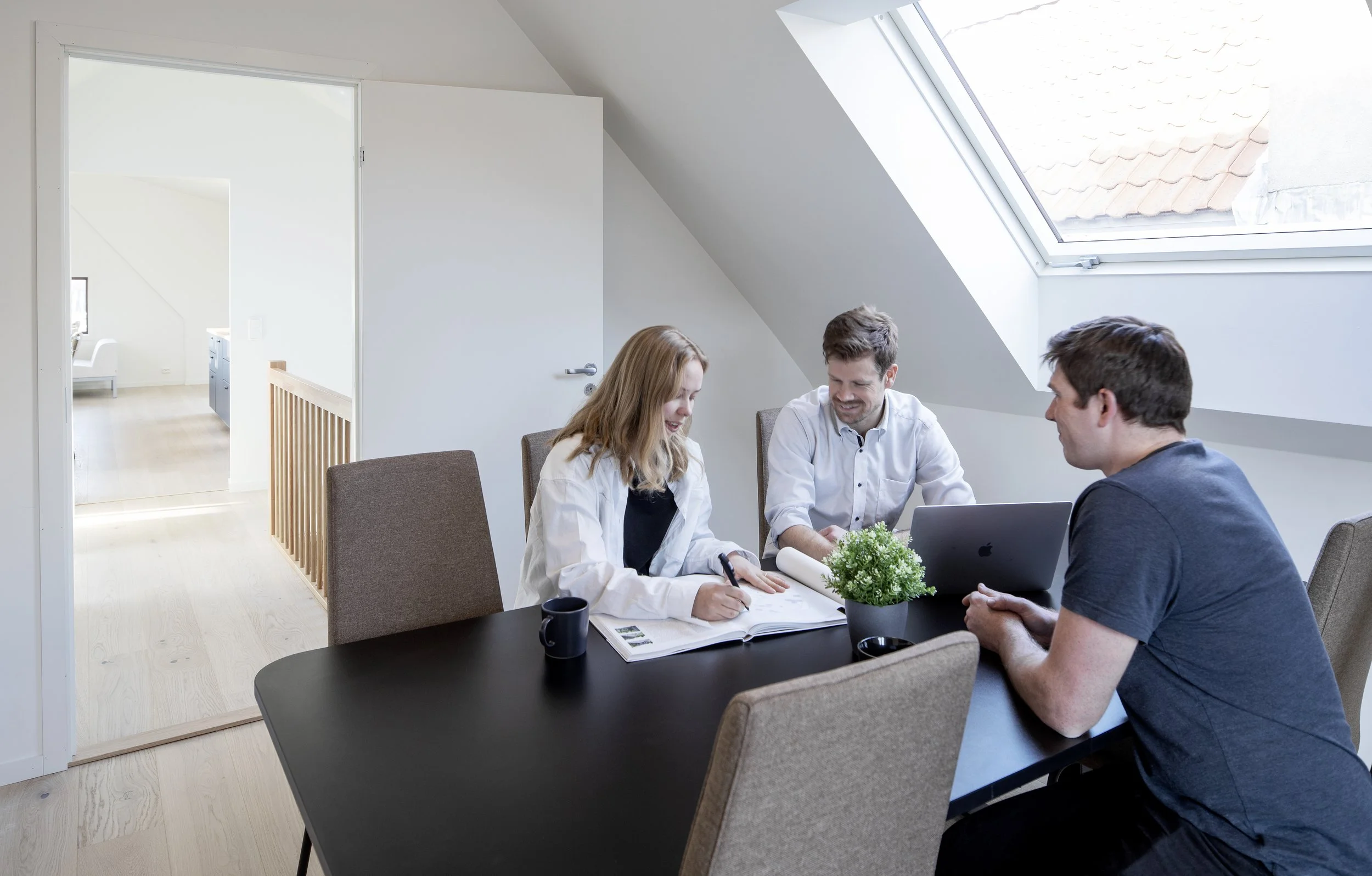 Tre personer diskuterer rundt et bord i et rom med skråtak og et stort vindu. De ser glade ut og har notatblokker og en bærbar PC på bordet.