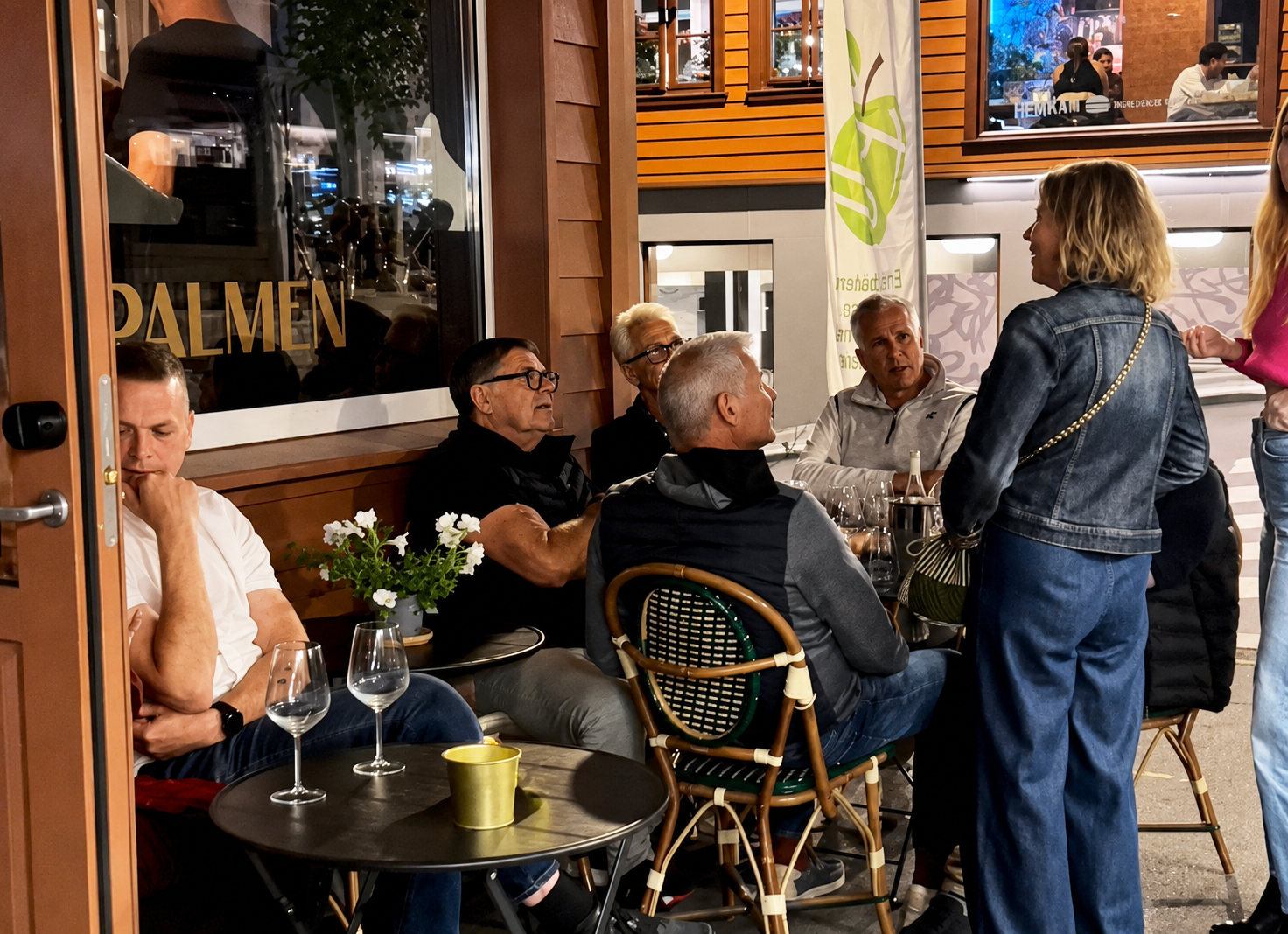 En gruppe mennesker sitter ved et bord utenfor en restaurant, de snakker med en kvinne som står. Det er flere tomme vinglass på bordet, og det ser ut til å være en uformell samtale.
