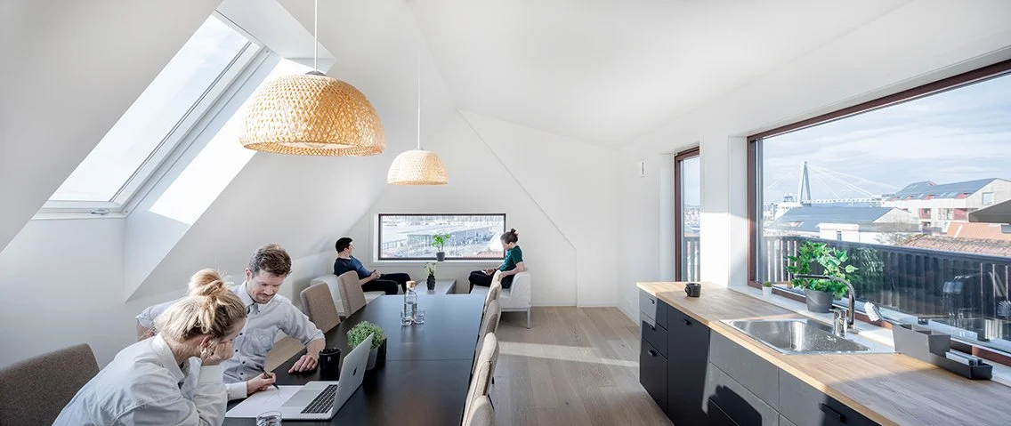 Moderne coworking-område med store vinduer, lys innendørs, grupper av mennesker som jobber ved bord, kjøkkenområde med vask og kjøkkenutstyr.