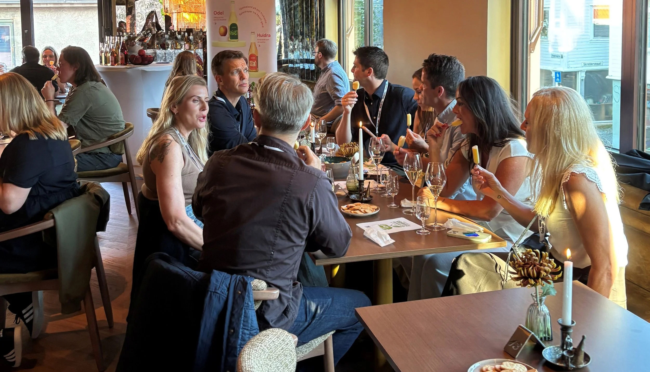 Mennesker sitter rundt et langbord i en restaurant og nyter dessert. Det er flere vin- og vannflasker på bordet, og noen har is på pinne. Noen personer snakker, og det er levende lys og blomster på bordet.