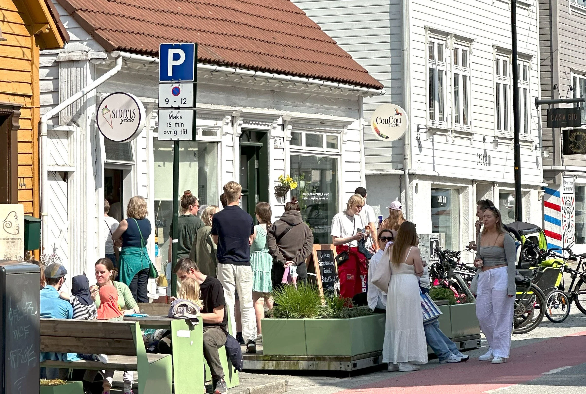 Grupp mennesker som står og sitter utendørs foran en hvit bygning med butikkfensere. Det er skilt som sier parkering og noe matrelatert reklame, og det er sykler og blomsterpotter. Mennesker er i samtale og ser ut til å nyte en dag ute.