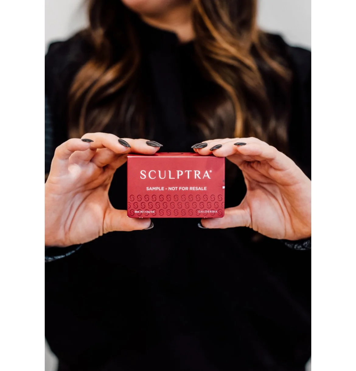 A woman with long brown hair and black nail polish holds a small red box of Sculptra sample in front of her chest.