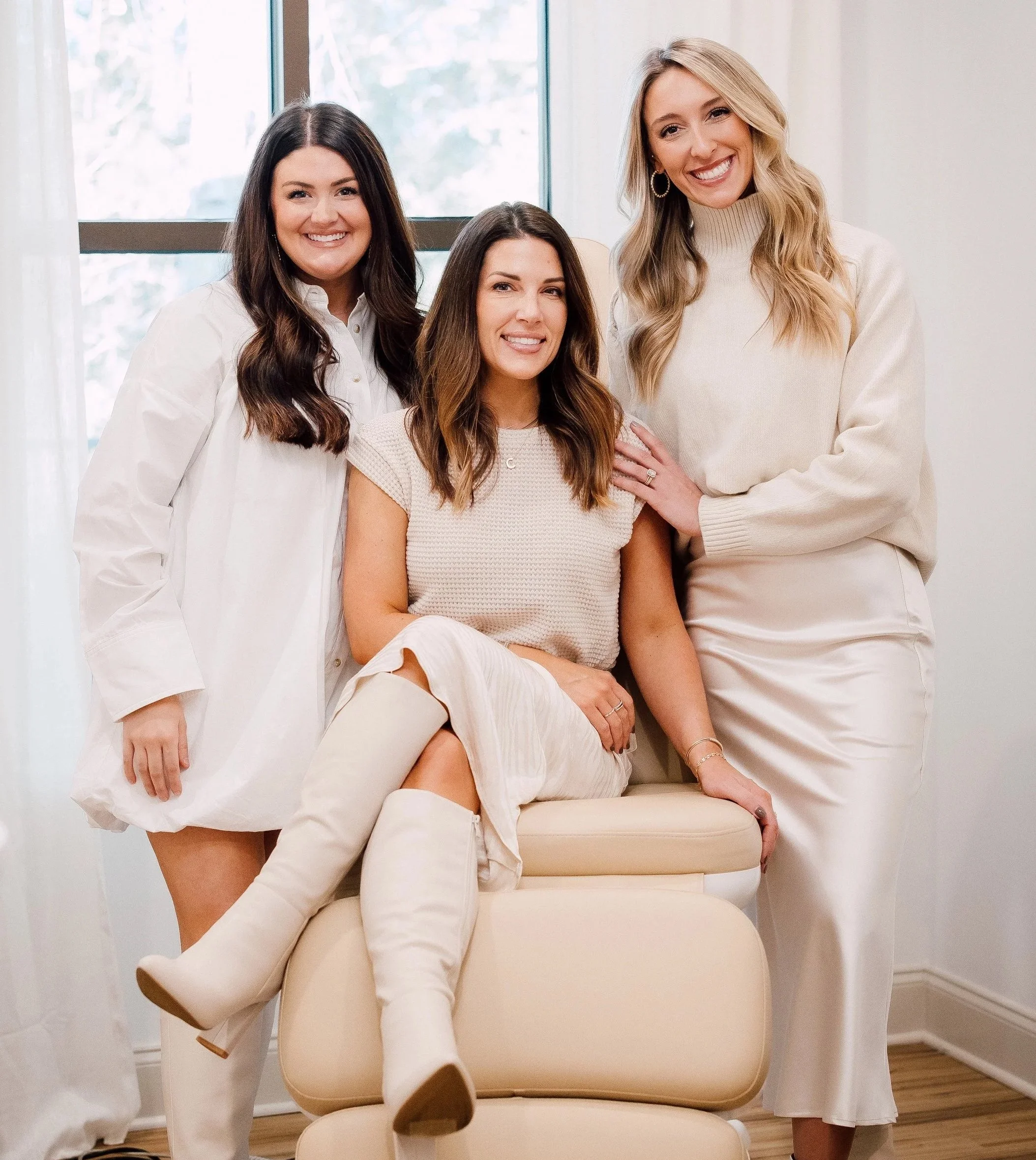 Three women posing together indoors, smiling, with a window in the background and a beige armchair in front of them.