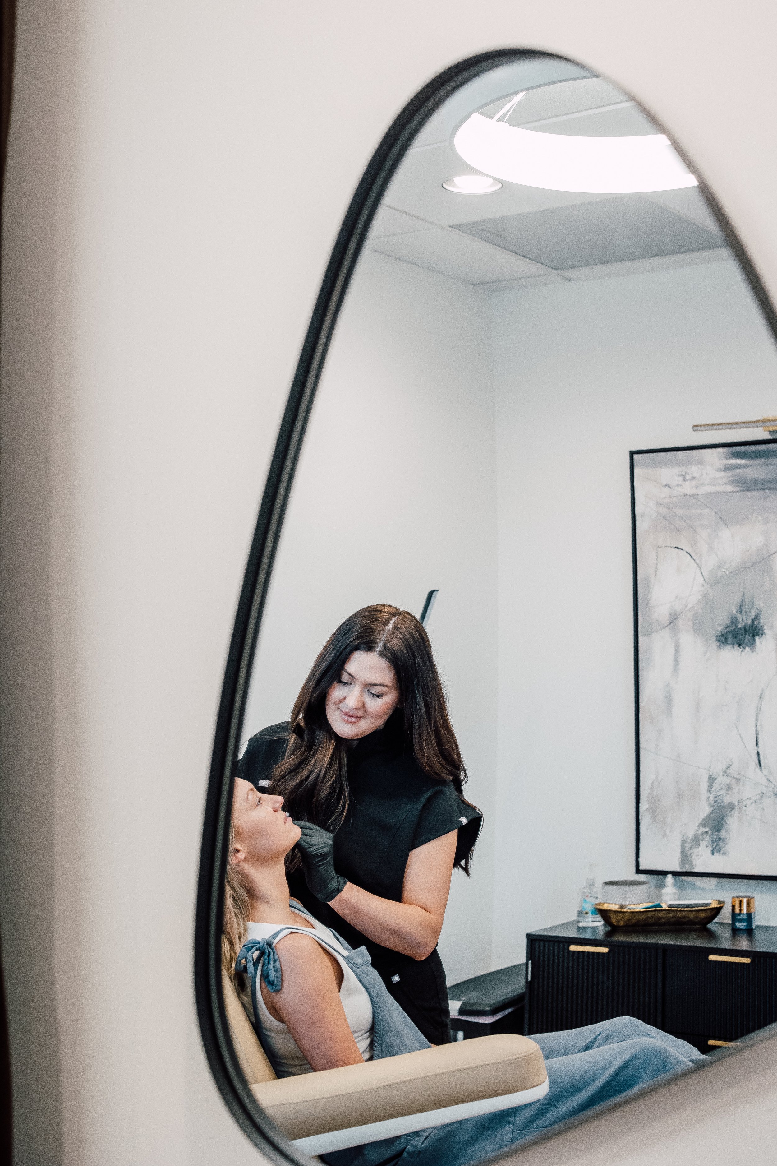 A woman is lying on a treatment bed while a skincare professional applies a cosmetic treatment to her face, observed through a mirror in a modern clinic room.