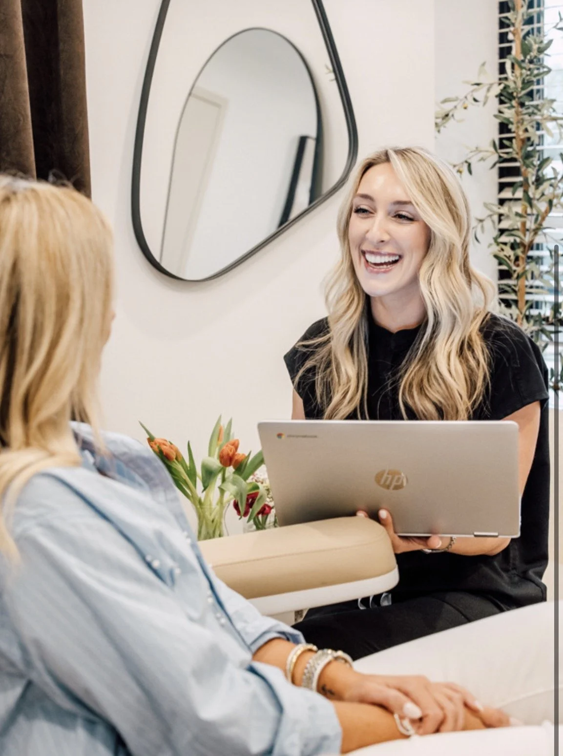 Two women having a conversation in a bright room, one holding a laptop, with a mirror and window in the background, and a vase of flowers on the table.