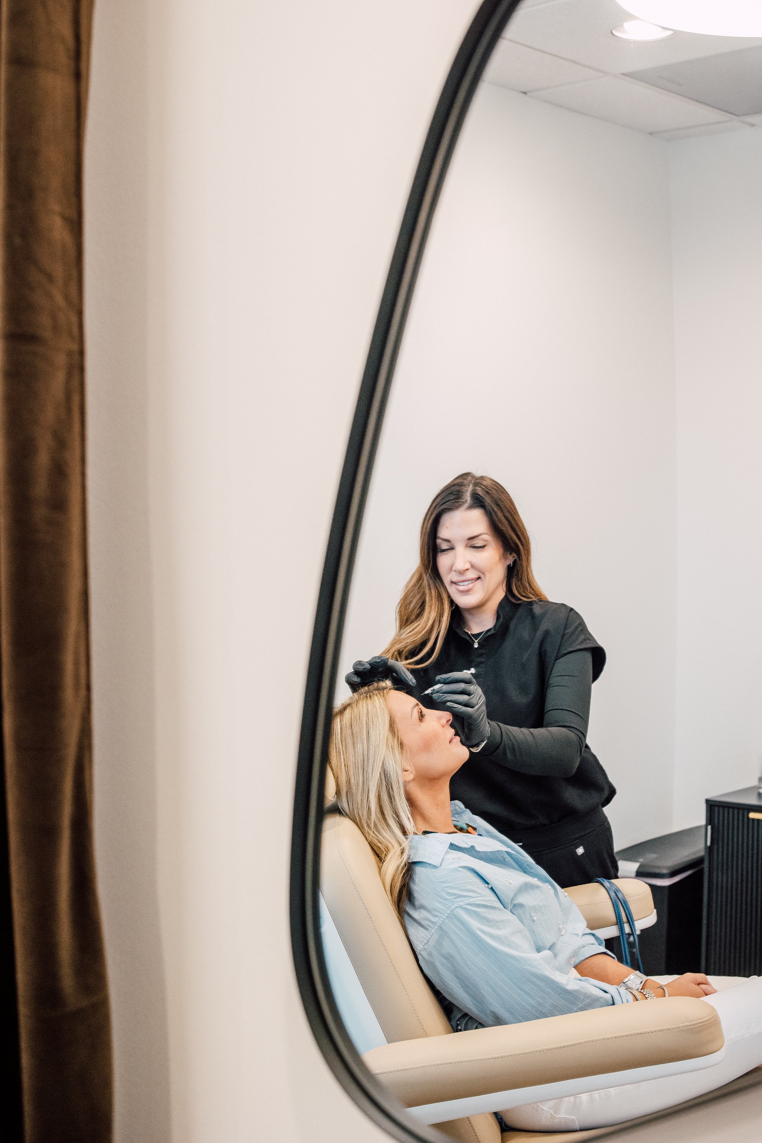 A woman with blonde hair and a blue striped shirt is seated in a salon chair, receiving a hair treatment from a stylist dressed in black, who is wearing black gloves and working on her hair. The scene is viewed through an oval mirror with a wooden frame.