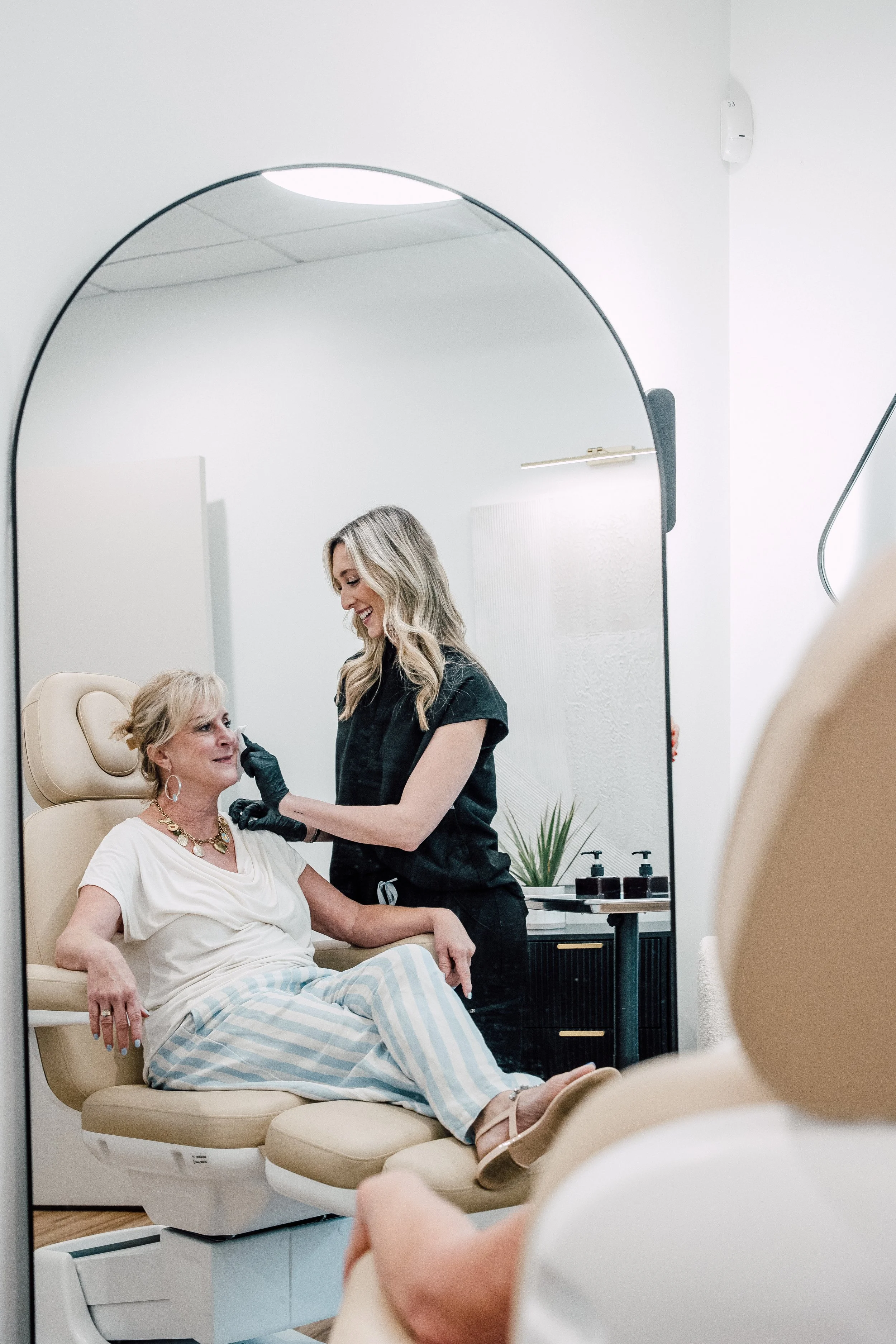 A woman with blonde hair and a white shirt receives a cosmetic procedure from a female technician with blonde hair in a black uniform. They are in a sterile clinic room with a mirror and supplies on a nearby table.