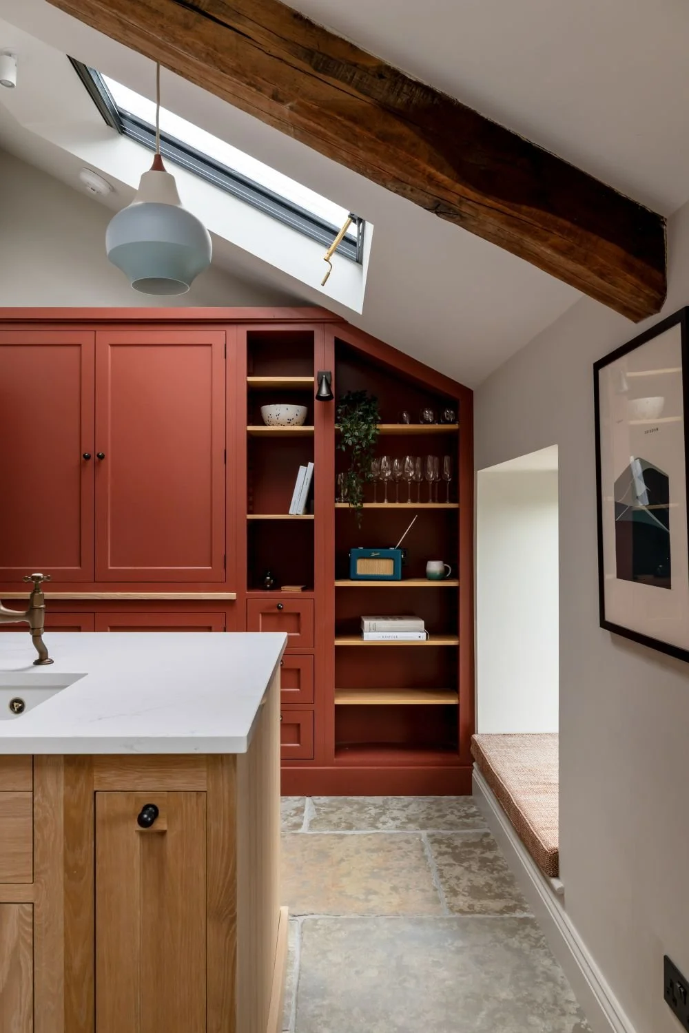 Interior of a home kitchen with a white countertop, wooden cabinetry, a skylight window, exposed wooden beam, and a built-in shelving unit with glasses and decorative items.