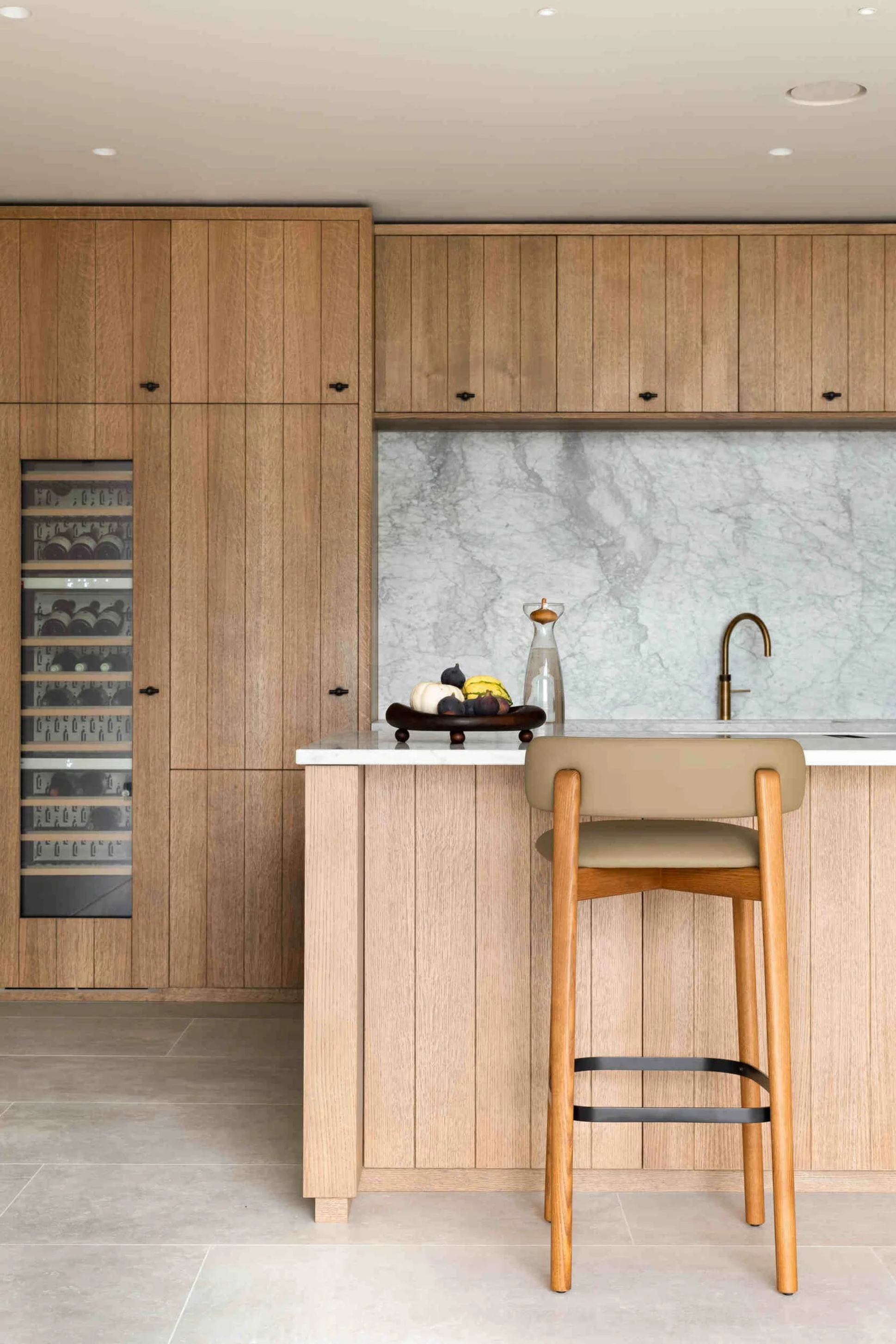 Modern kitchen interior with light wood cabinets, a marble backsplash, a sink with a brass faucet, and a beige chair at a kitchen island. Decorative pumpkins and a vase on the counter.