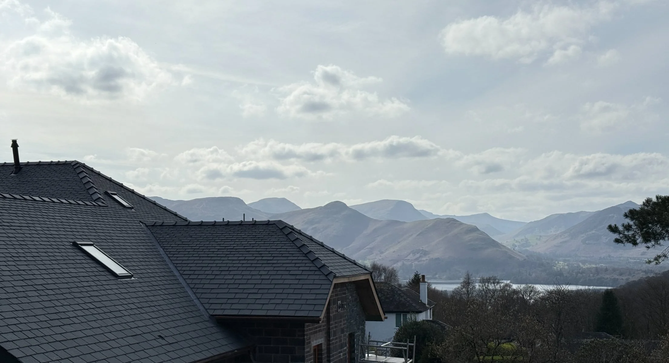 Roof with dark gray shingles and skylights, mountains in the background under a cloudy sky.