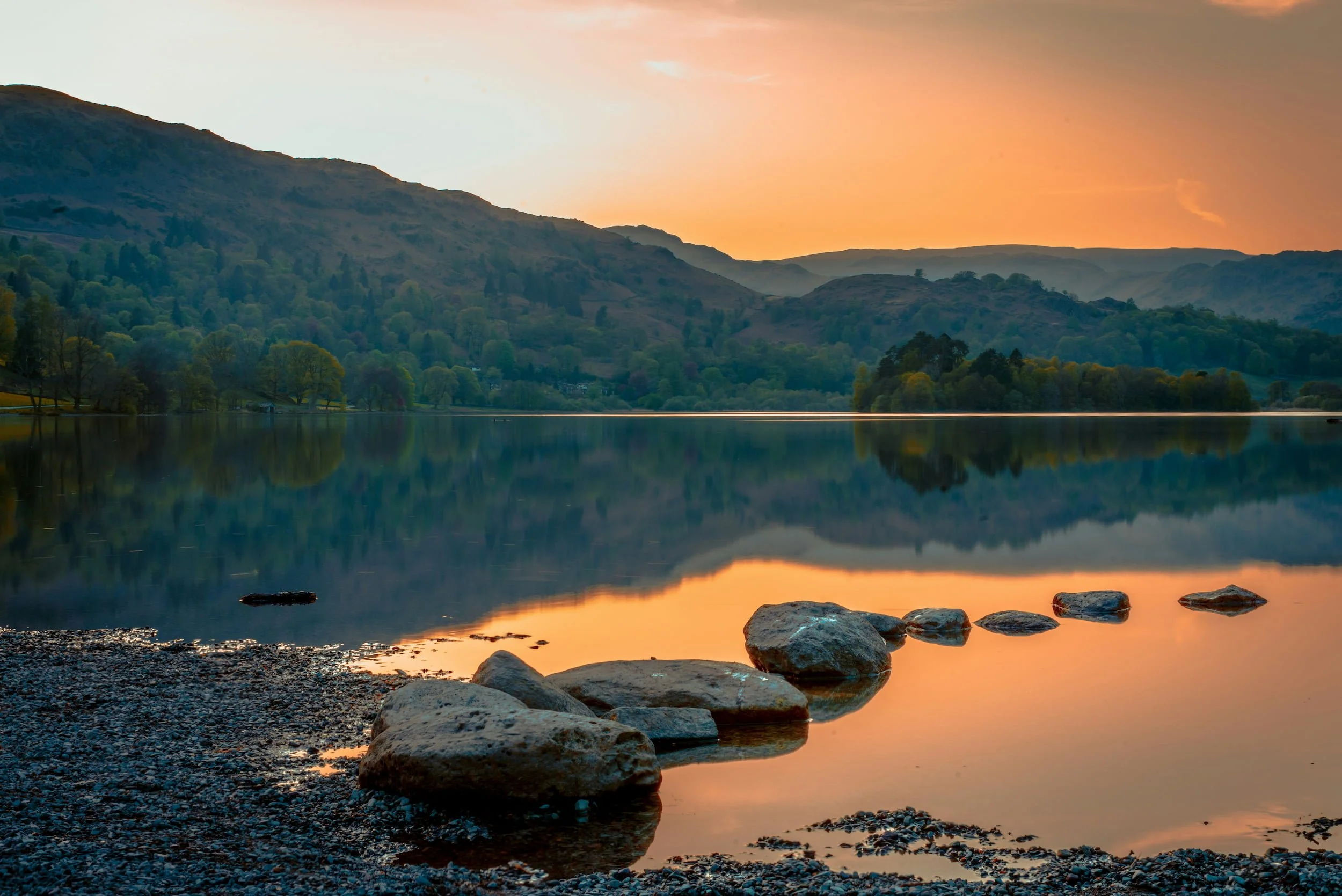 Scenic view of a calm lake at sunset, surrounded by green hills and mountains, with rocks along the shoreline and a colorful sky reflected in the water.