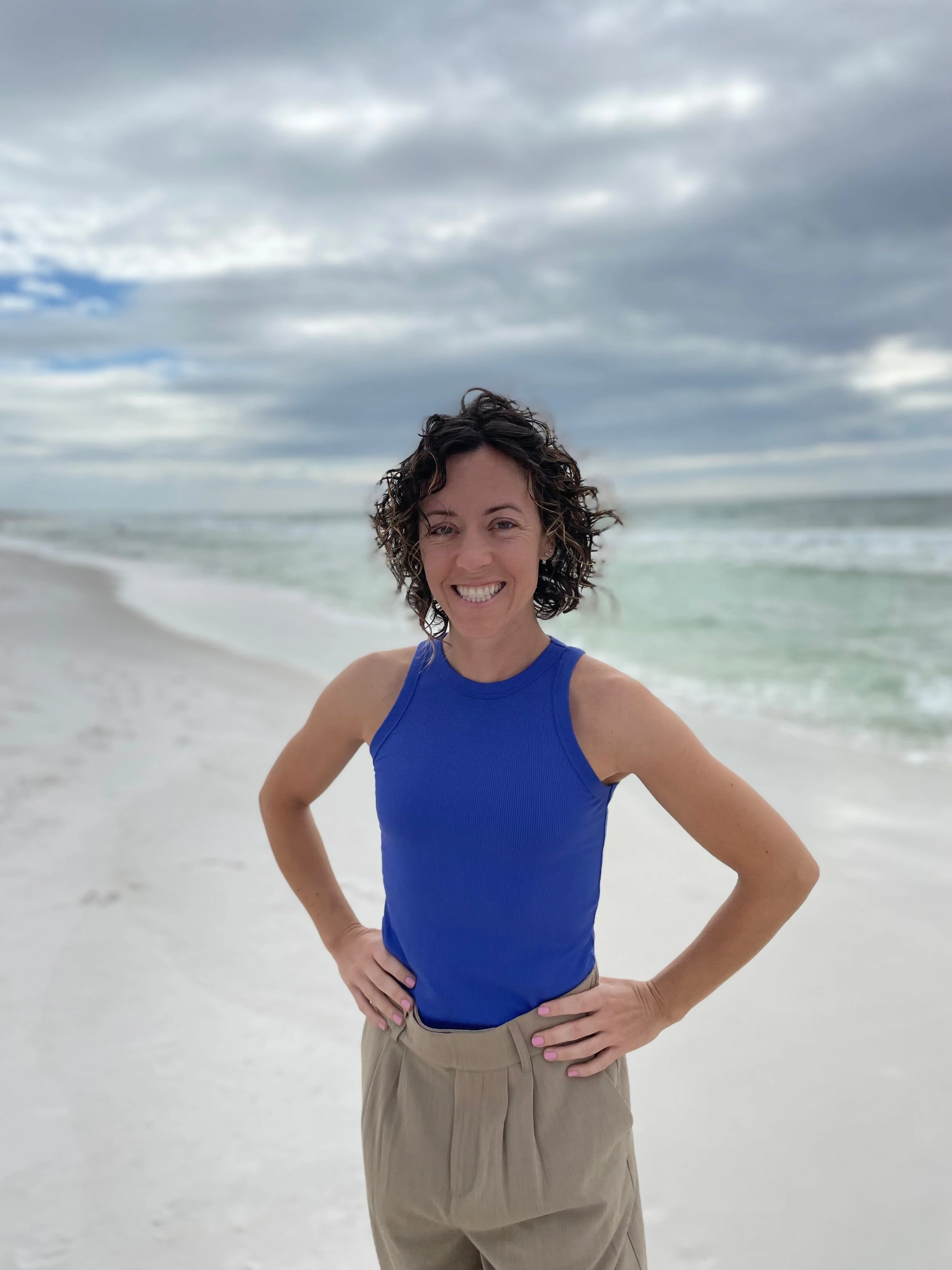 A woman with curly hair wearing a blue sleeveless top and beige pants stands on a beach with white sand, smiling at the camera, with ocean waves and a cloudy sky in the background.