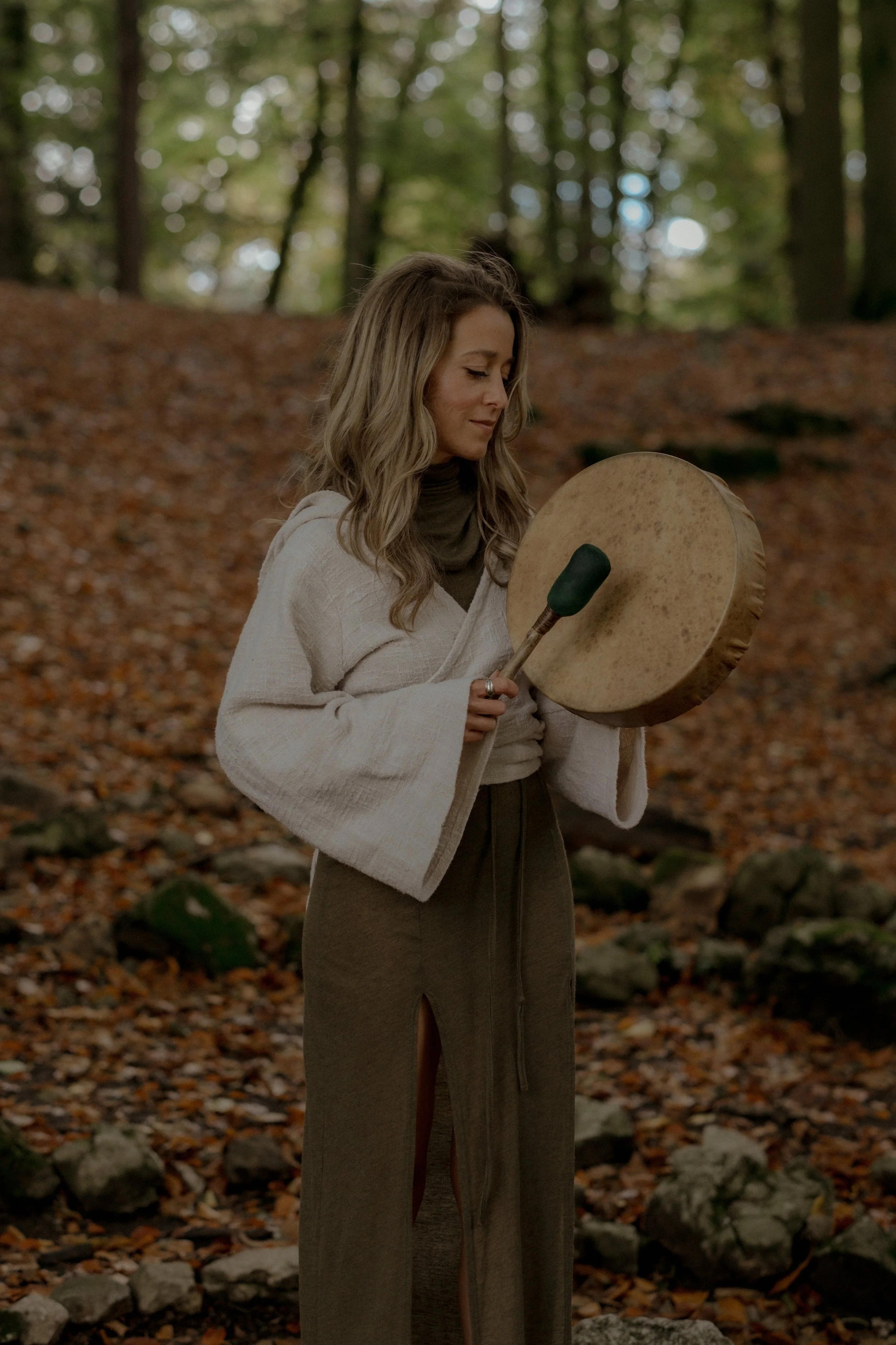 Woman in a beige sweater and brown pants holding a percussion instrument outdoors in a forest with autumn leaves.