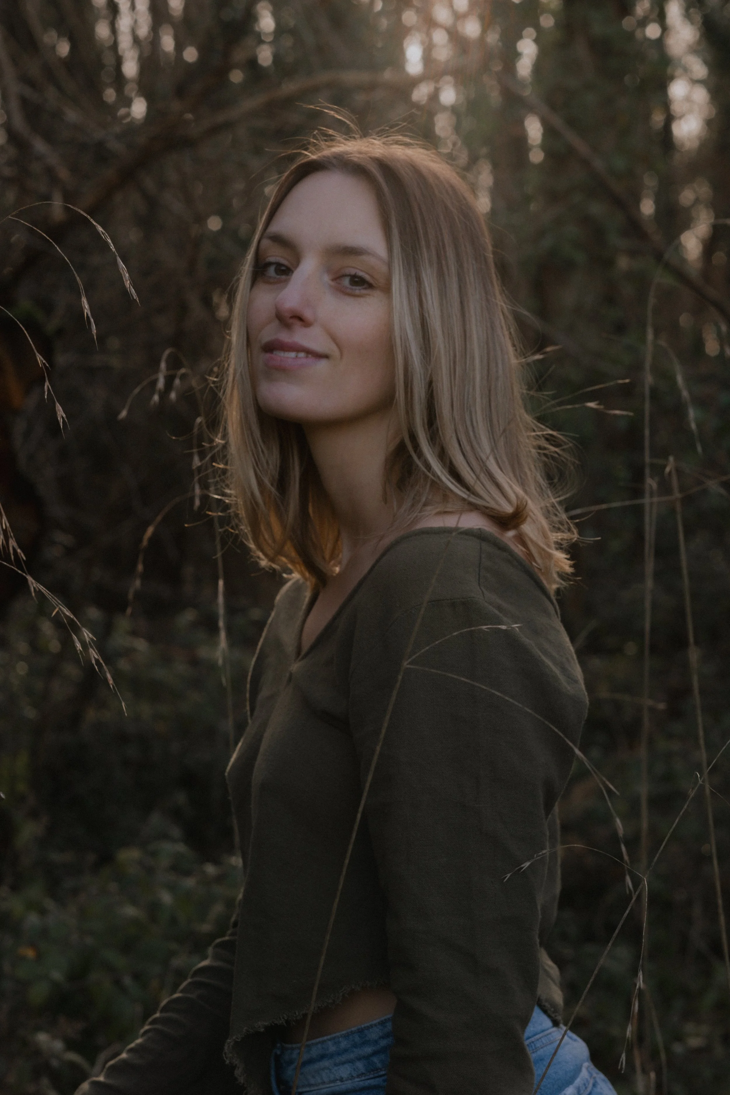 A young woman with shoulder-length blond hair, smiling slightly, standing outdoors in a wooded area with trees in the background during late afternoon or early evening.