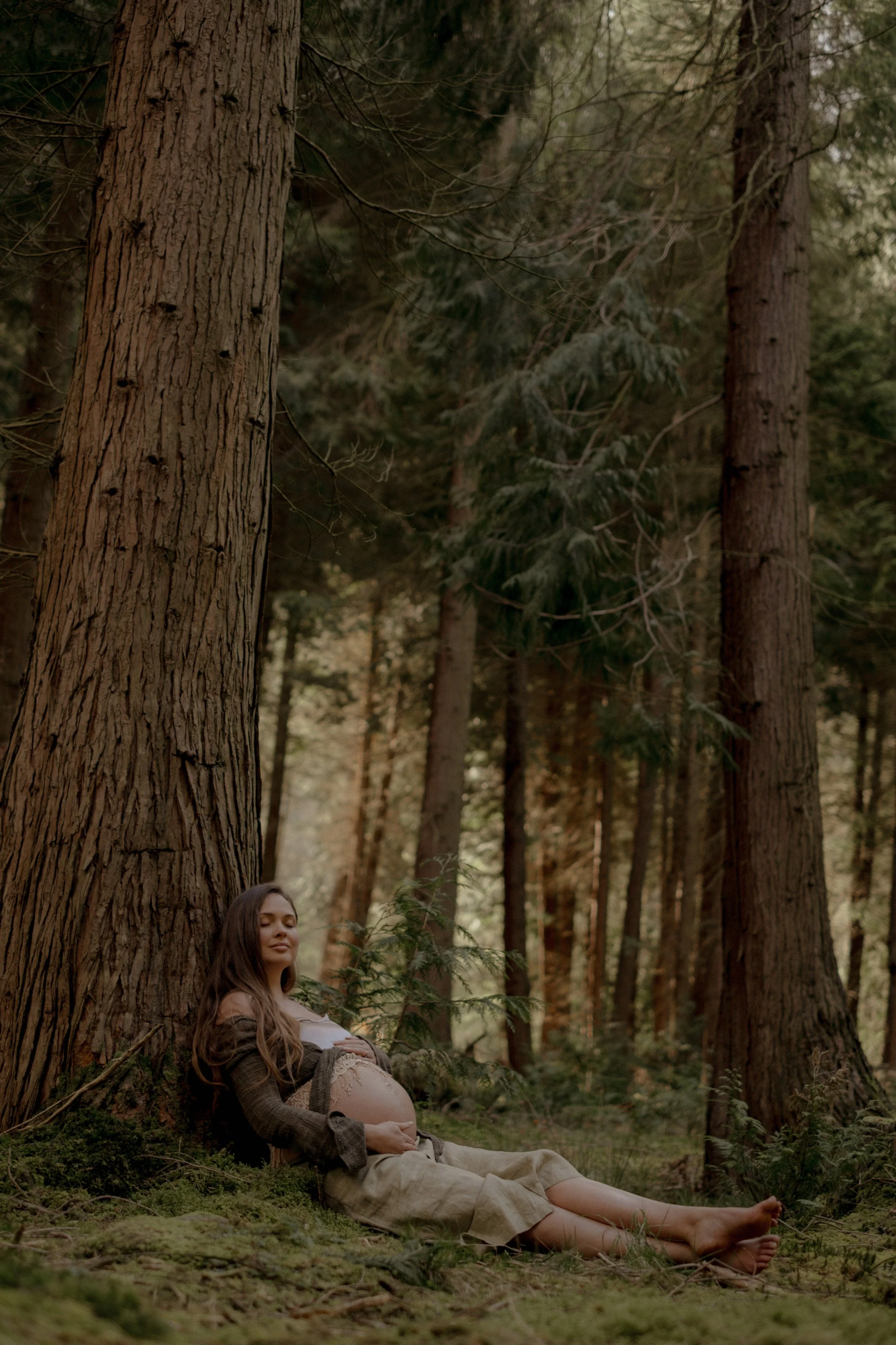 Mother leaning against a tree in a forest during a relaxed, natural motherhood maternity outdoor photoshoot in Wales