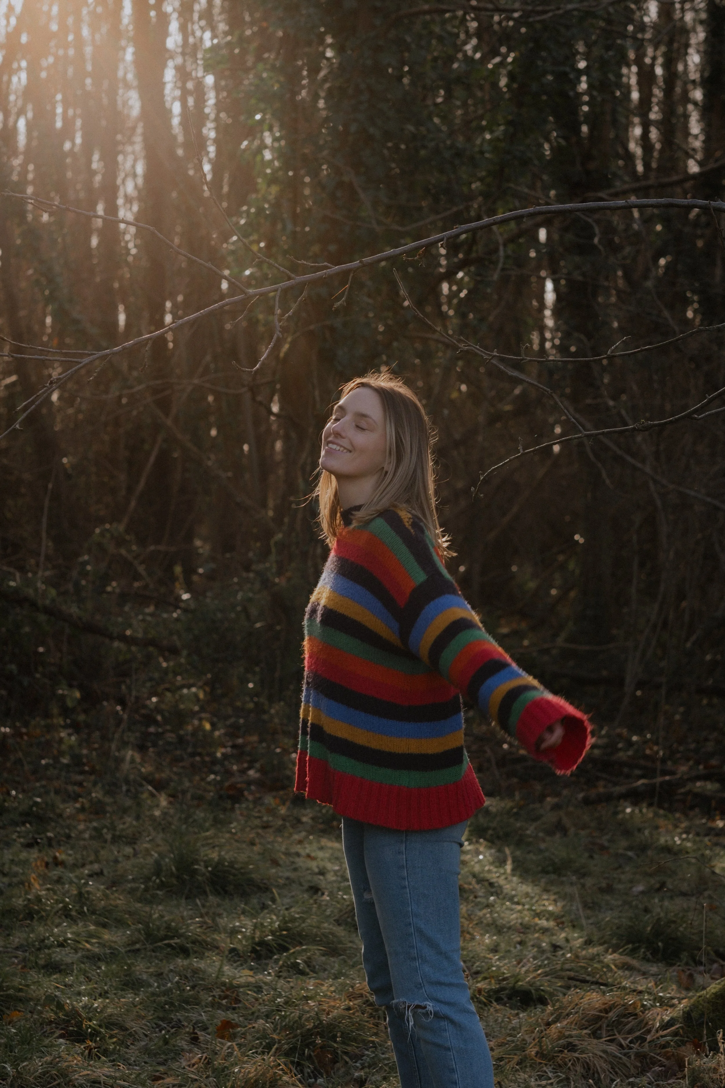 A young woman outdoors in a forest, smiling with eyes closed, wearing a colorful striped sweater and jeans, with sunlight filtering through trees.