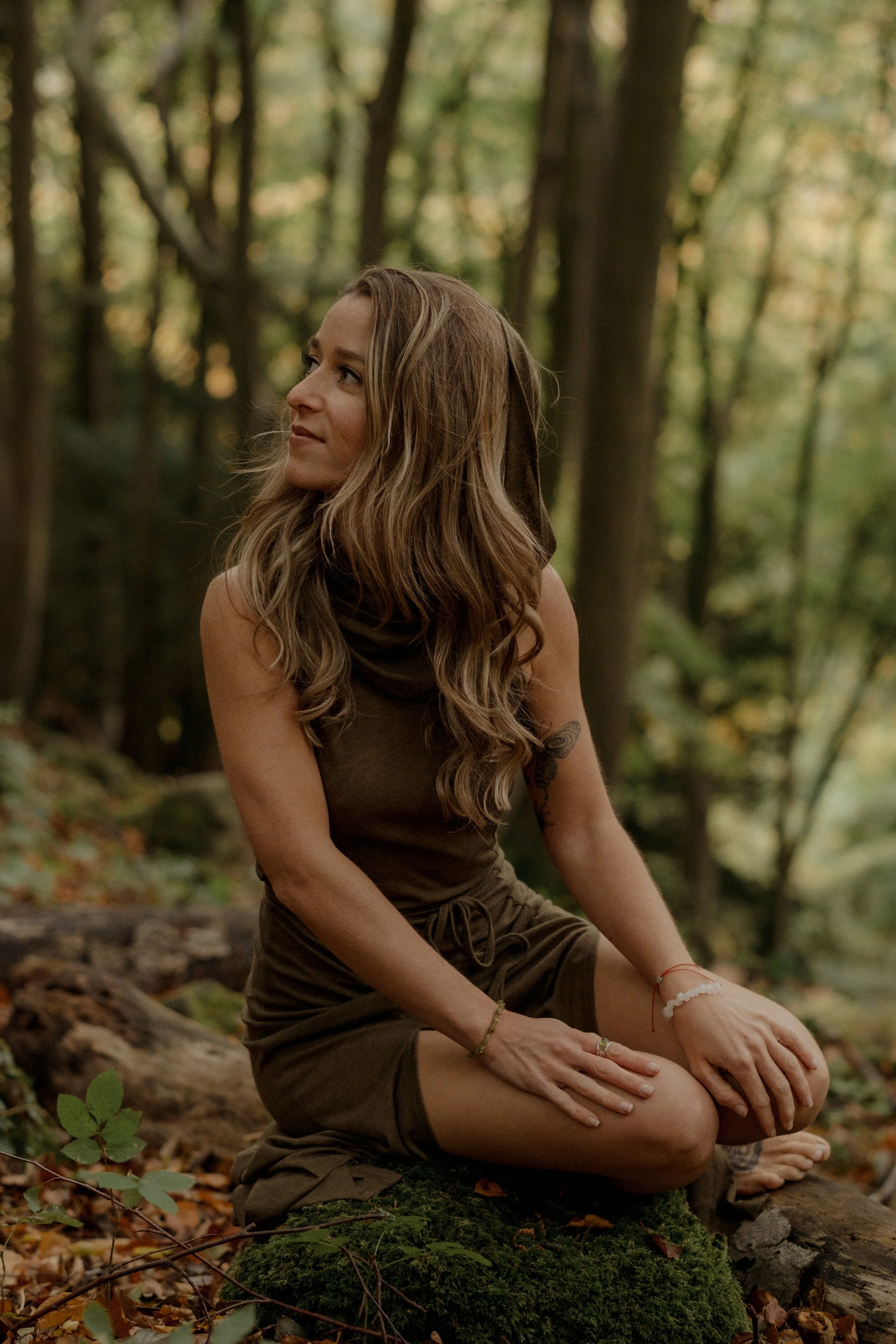 A woman with long wavy hair sitting cross-legged on a mossy log in a forest, dressed in a brown sleeveless dress, surrounded by trees and autumn leaves.