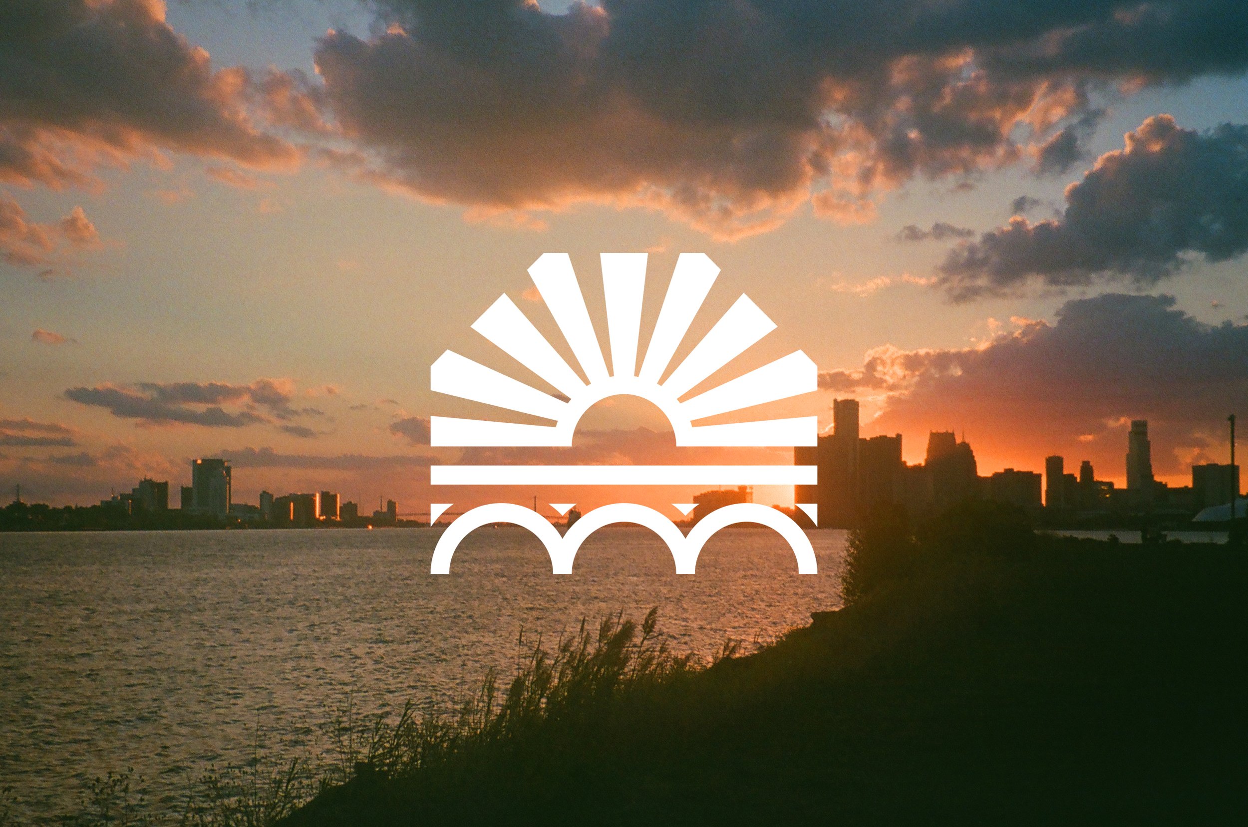 Sunset over a city skyline with a water body in the foreground, and a white graphic of a rising sun above an arched bridge overlaying the scene.