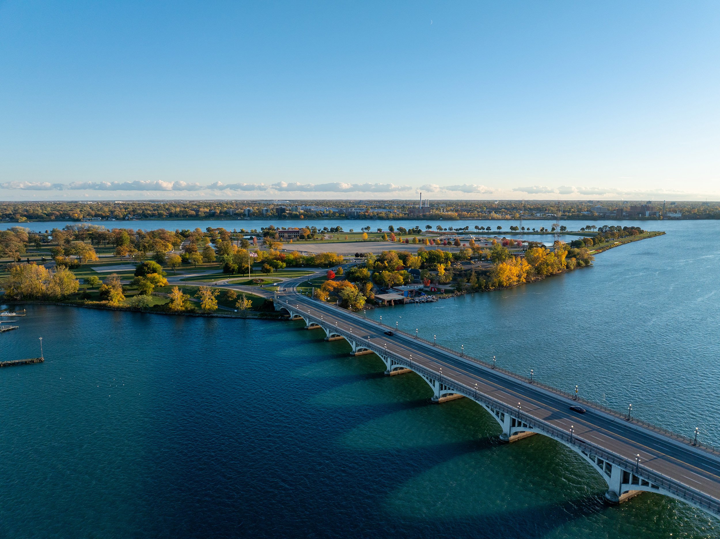 An aerial view of a bridge crossing a large body of water, with trees showing fall colors, and a park and city skyline in the background.